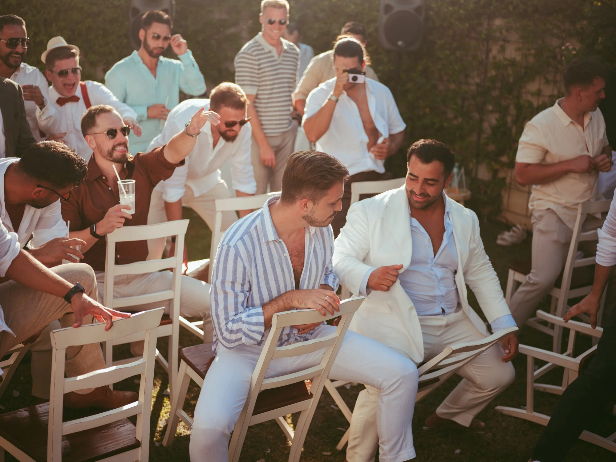 Lo sposo in abito bianco e gli invitati uomini seduti in cerchio sulle sedie durante un gioco goliardico al matrimonio