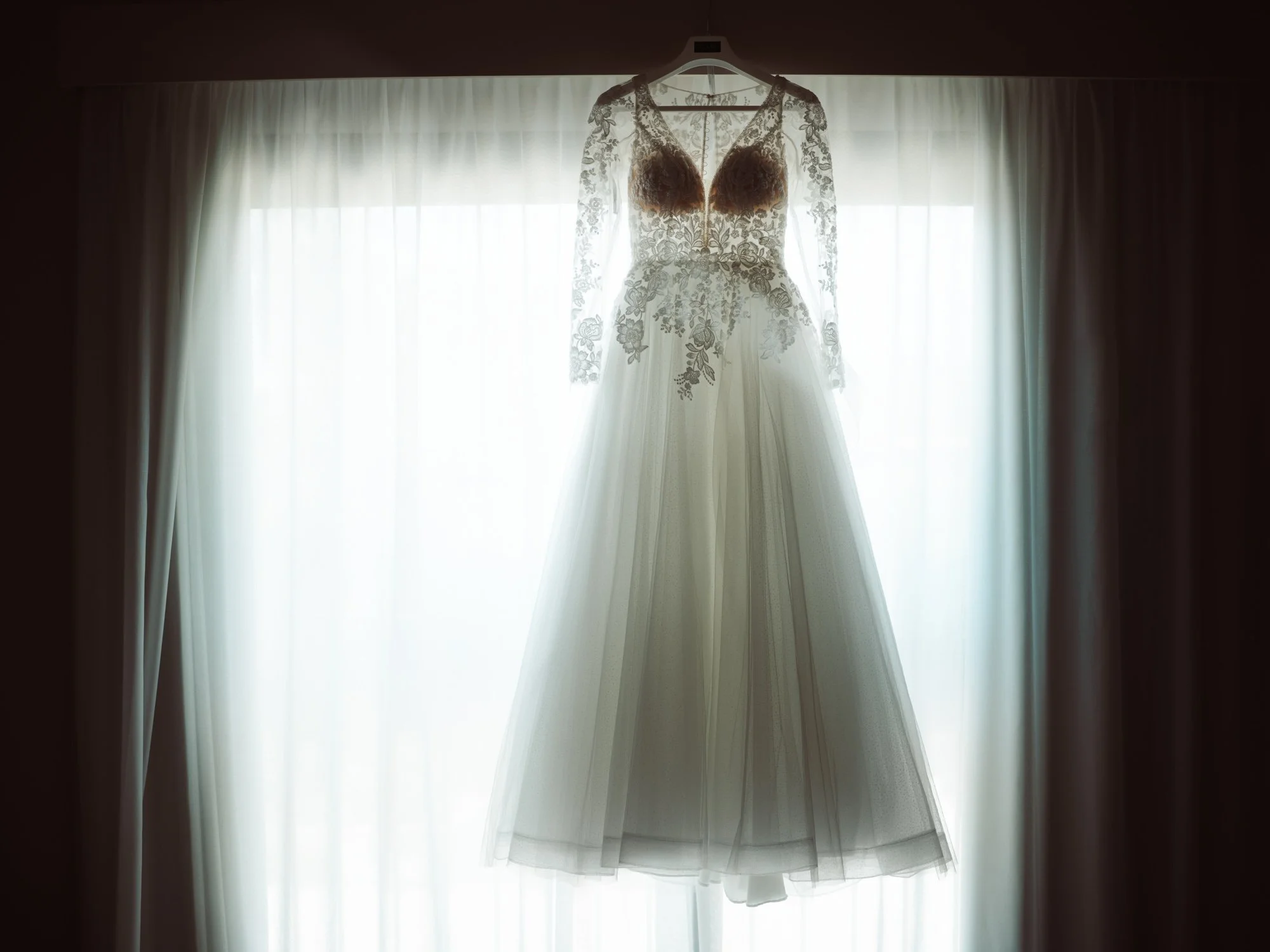 Abito da sposa in pizzo con maniche lunghe e gonna in tulle appeso davanti a una finestra illuminata