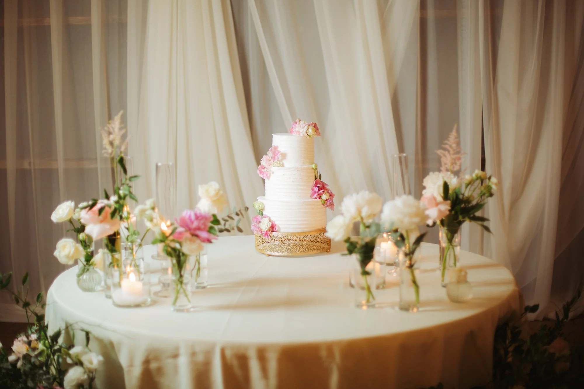 Torta nuziale a tre piani decorata con ortensie e peonie su un tavolo tondo circondato da vasi e candele
