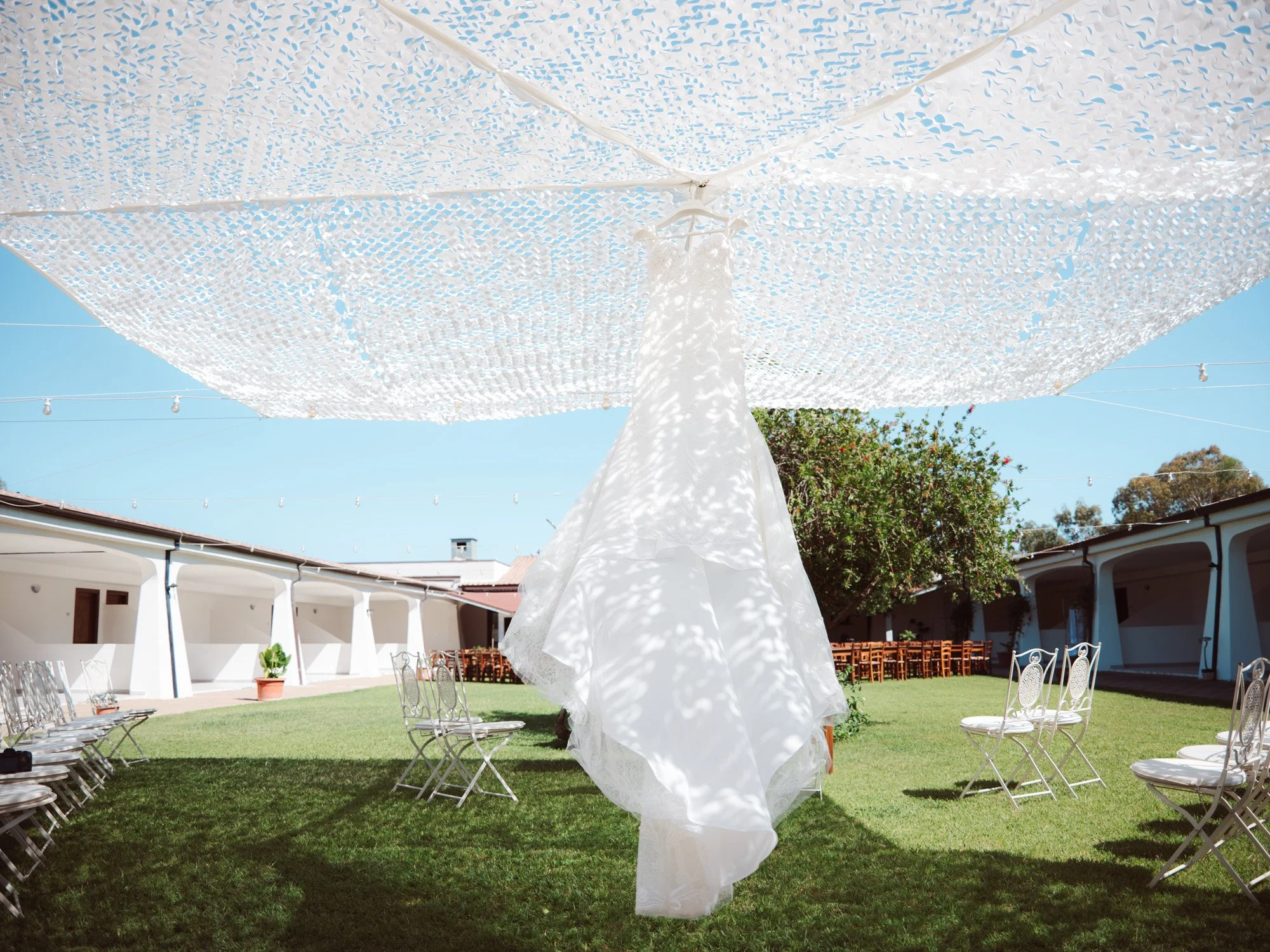 L'abito da sposa in pizzo sospeso nel giardino di una location esclusiva in Sardegna, illuminato dalla luce del sole.