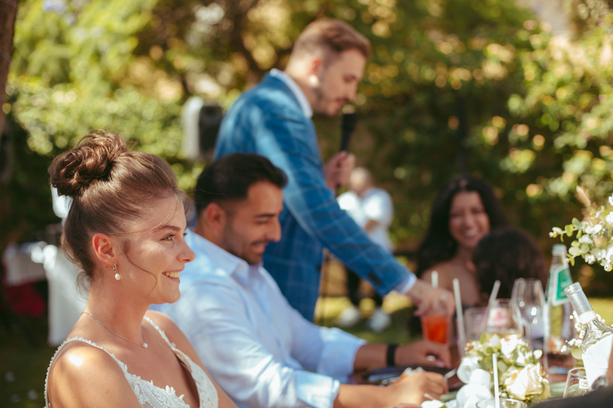 Sposa e sposo seduti vicini sorridono felici mentre l'amico dello sposo parla al microfono in sottofondo