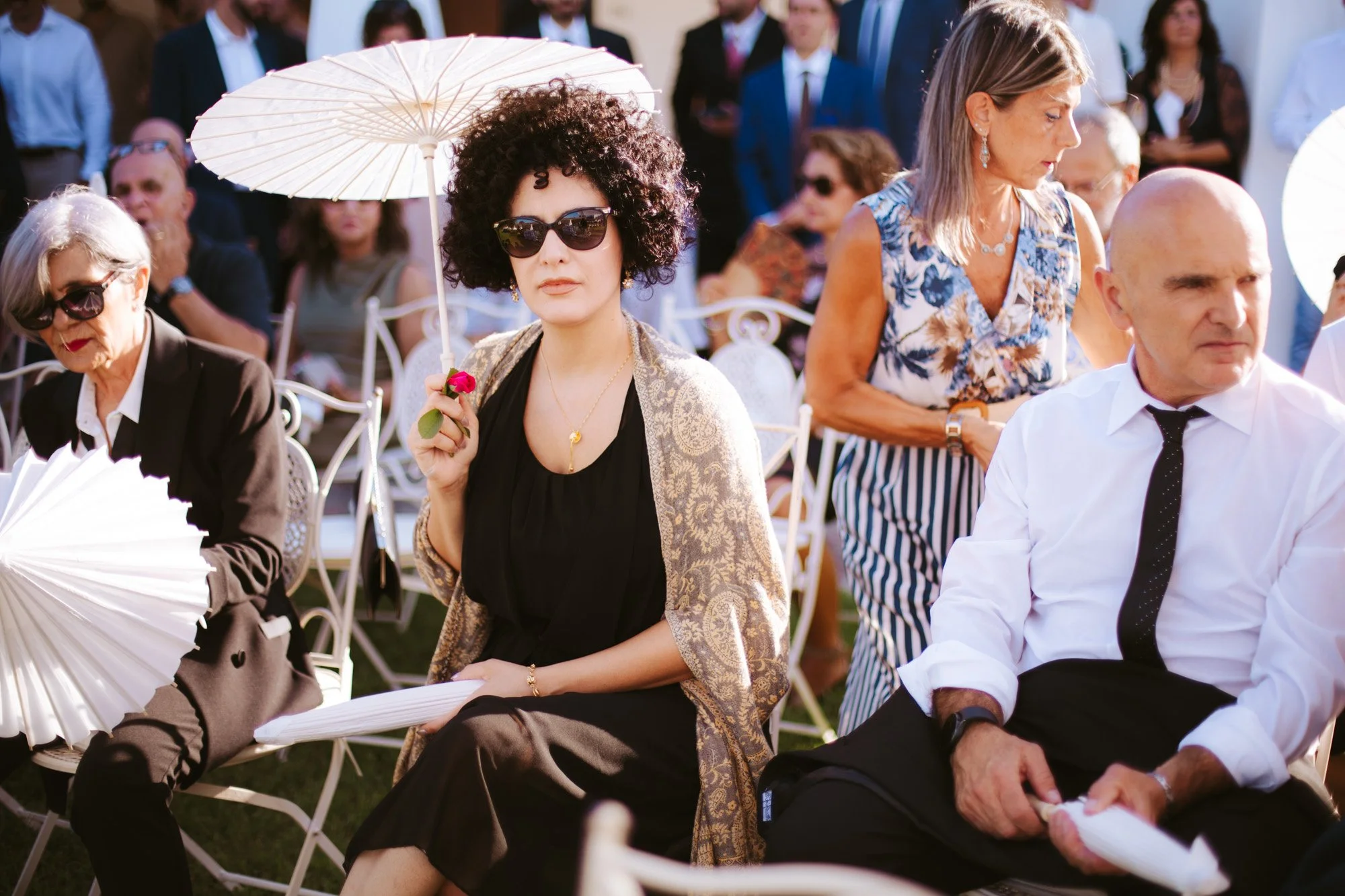 Eleganti invitati seduti tra il pubblico con ombrellini parasole durante la celebrazione del matrimonio.