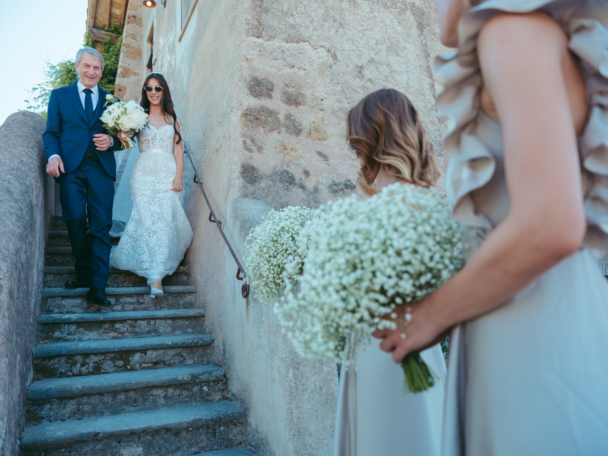 La sposa con occhiali da sole e bouquet scende la scalinata in pietra a braccetto con il padre
