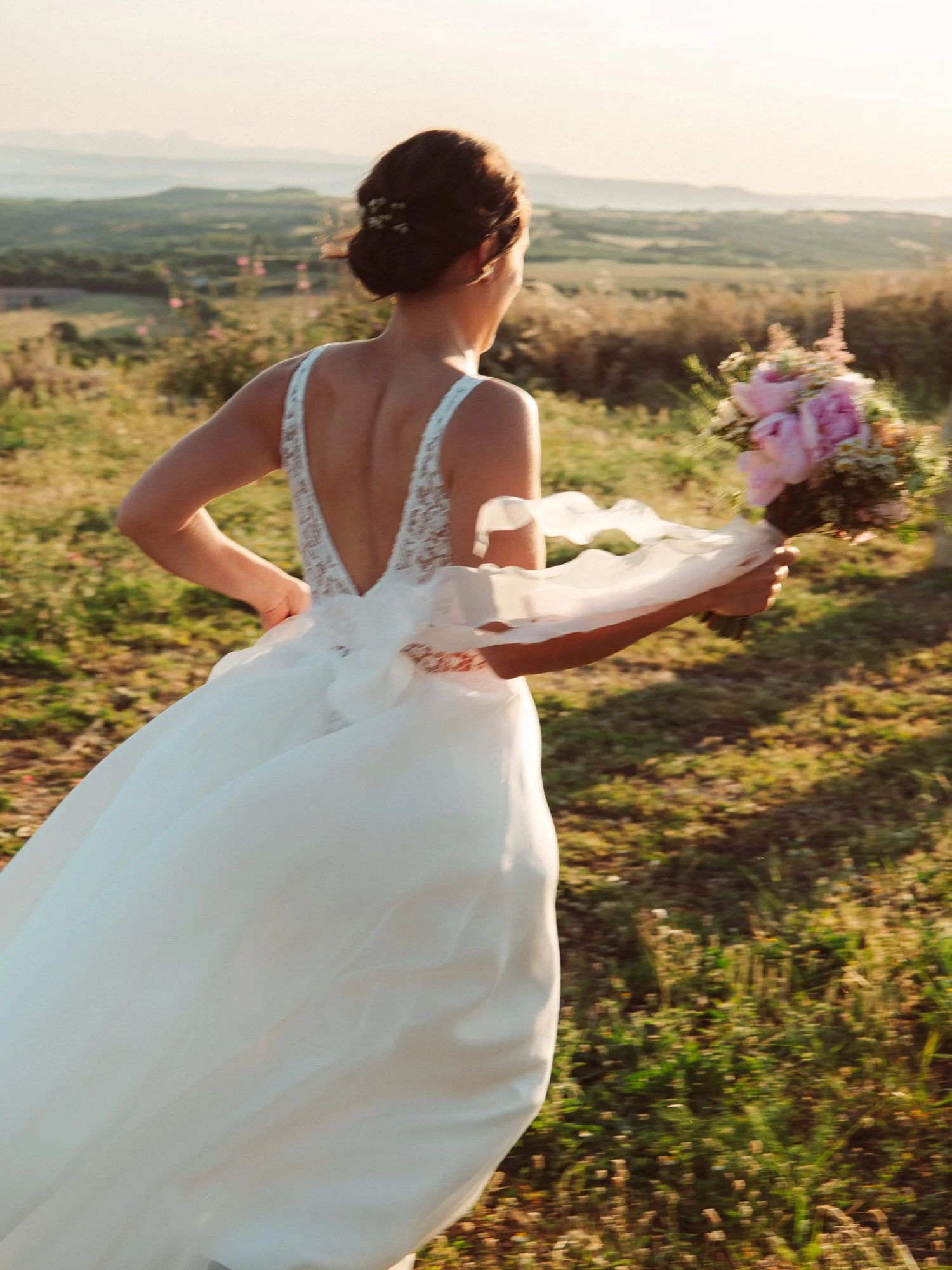 Sposa di spalle che corre nei campi tenendo il bouquet da sposa