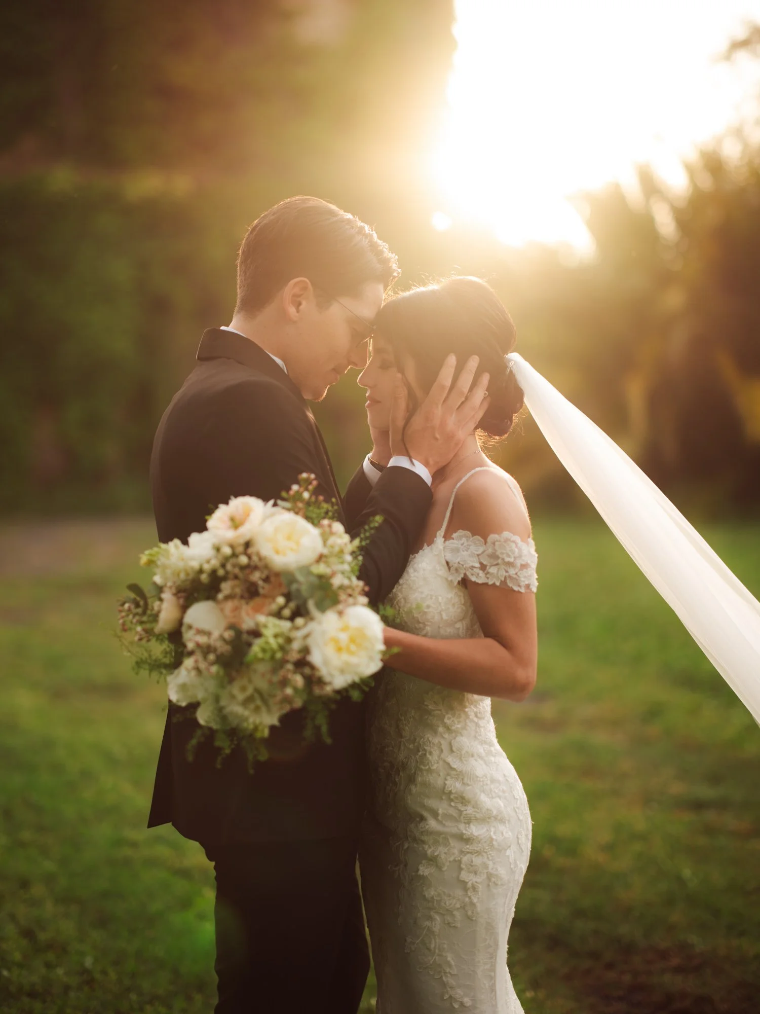 Sposi eleganti che si baciano sul prato al tramonto con il velo della sposa disteso sull'erba