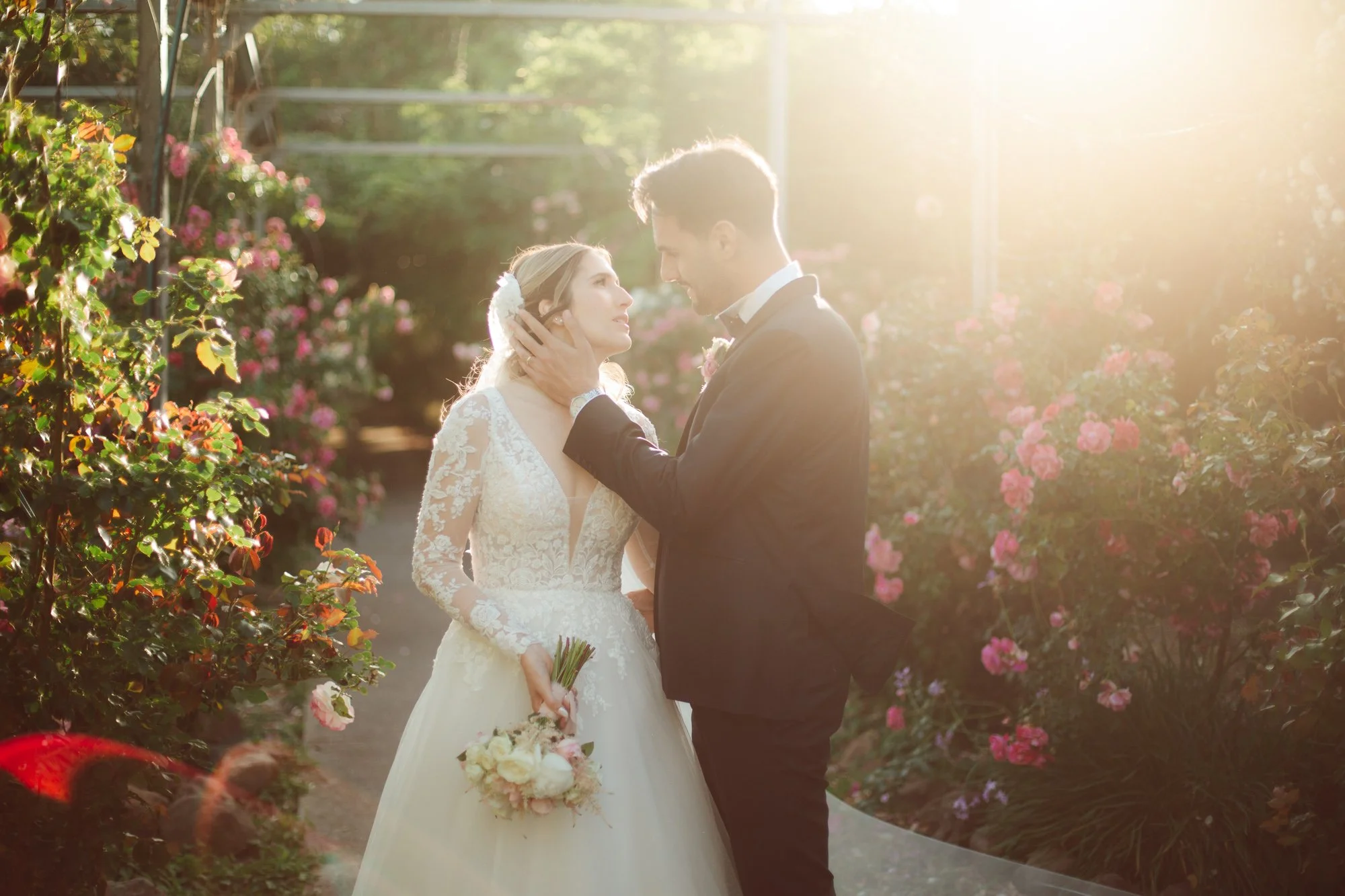 Lo sposo tiene delicatamente il viso della sposa tra le mani mentre si guardano immersi tra le rose