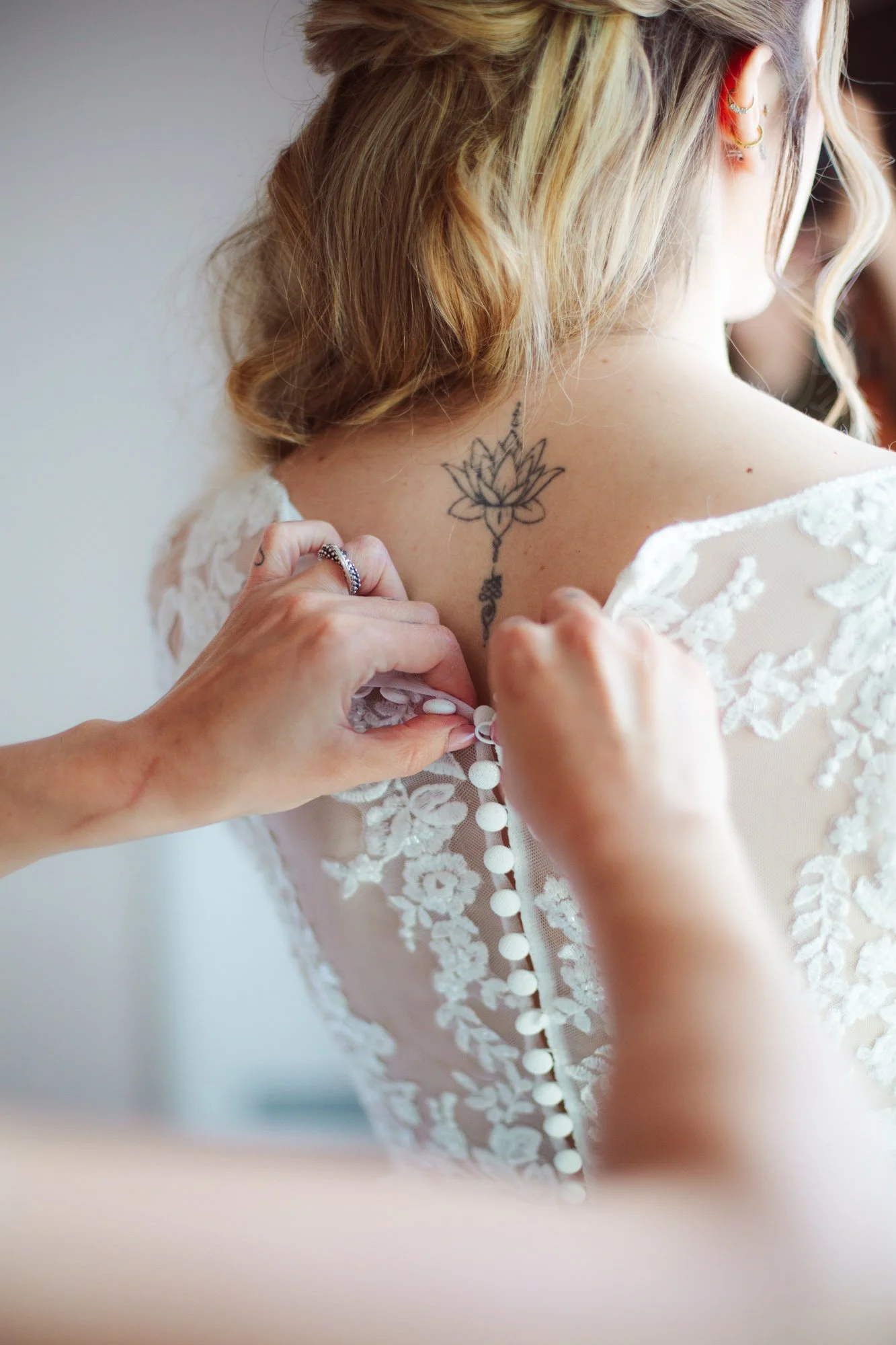 Mani che chiudono la fila di bottoncini dell'abito da sposa in pizzo sulla schiena con tatuaggio fiore di loto
