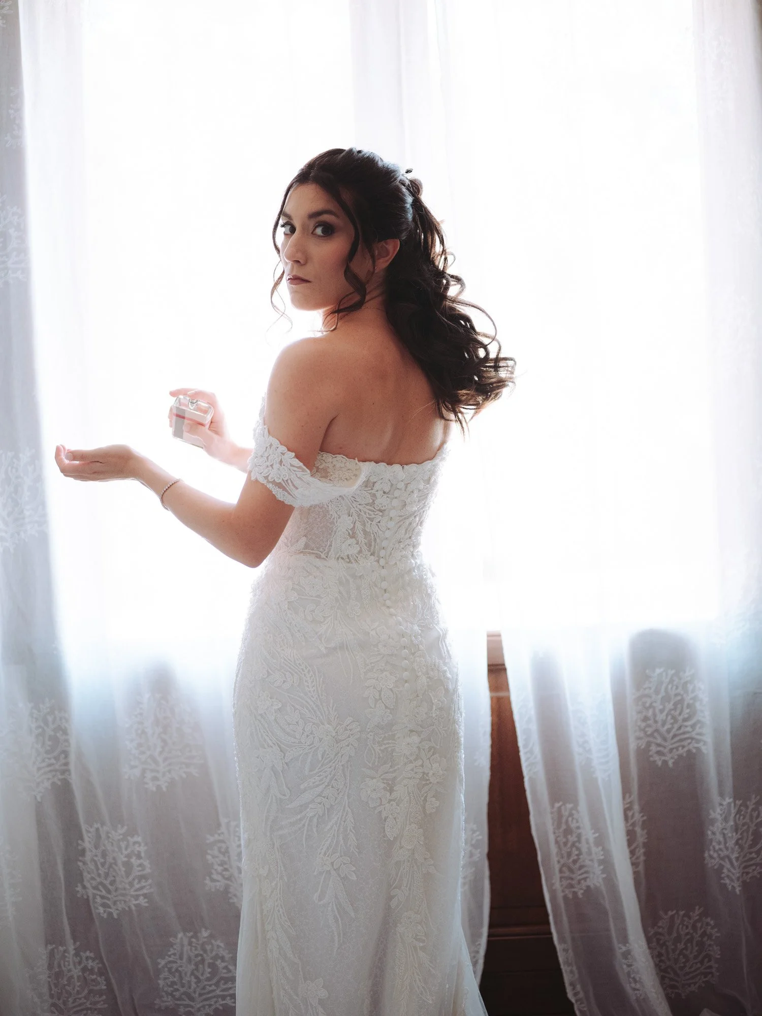La sposa indossa il profumo vicino alla finestra con luce naturale filtrata dalle tende durante i preparativi del matrimonio.