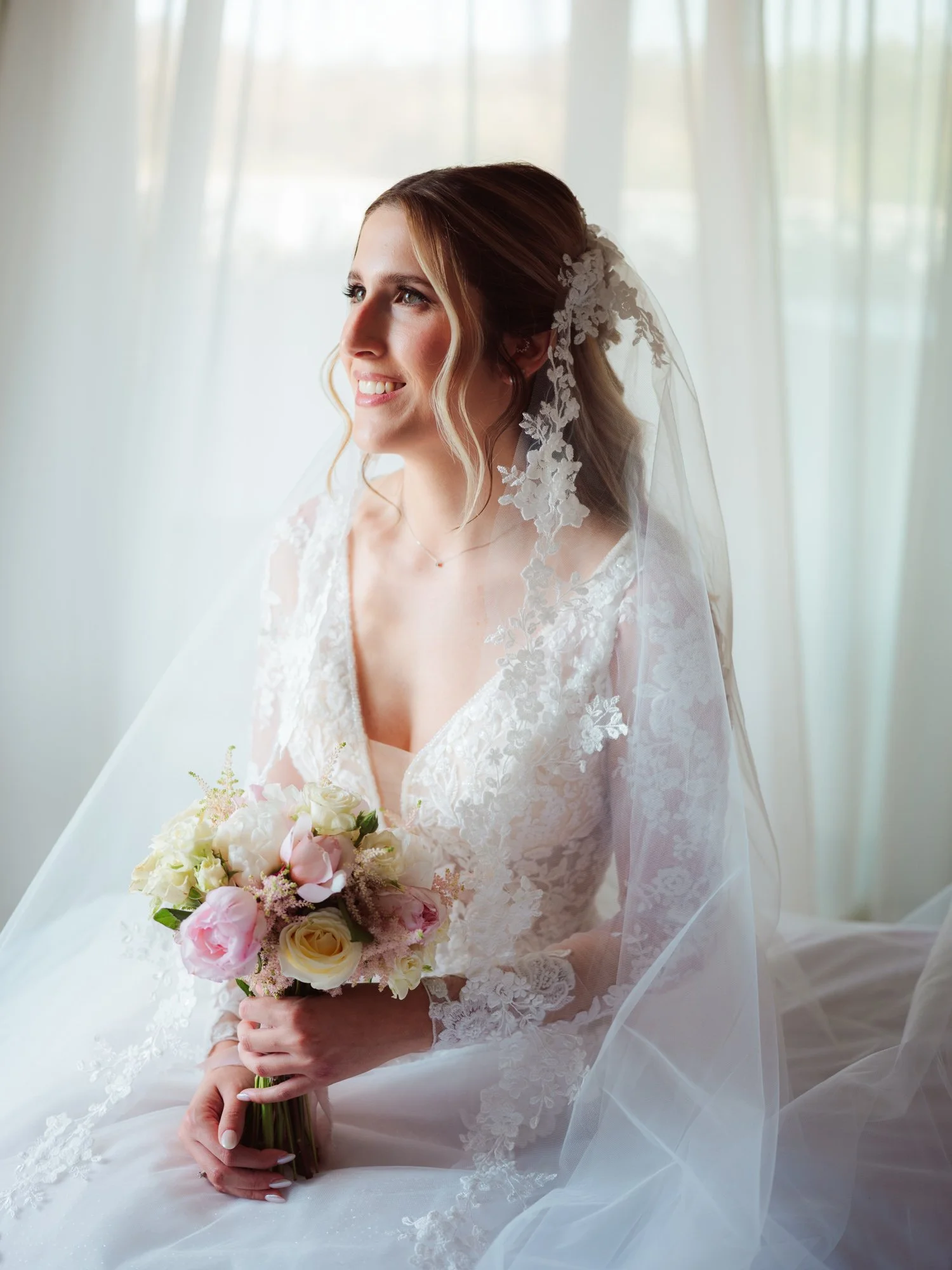 Sposa sorridente in abito da sposa in pizzo seduta accanto alla finestra mentre tiene il bouquet tra le mani