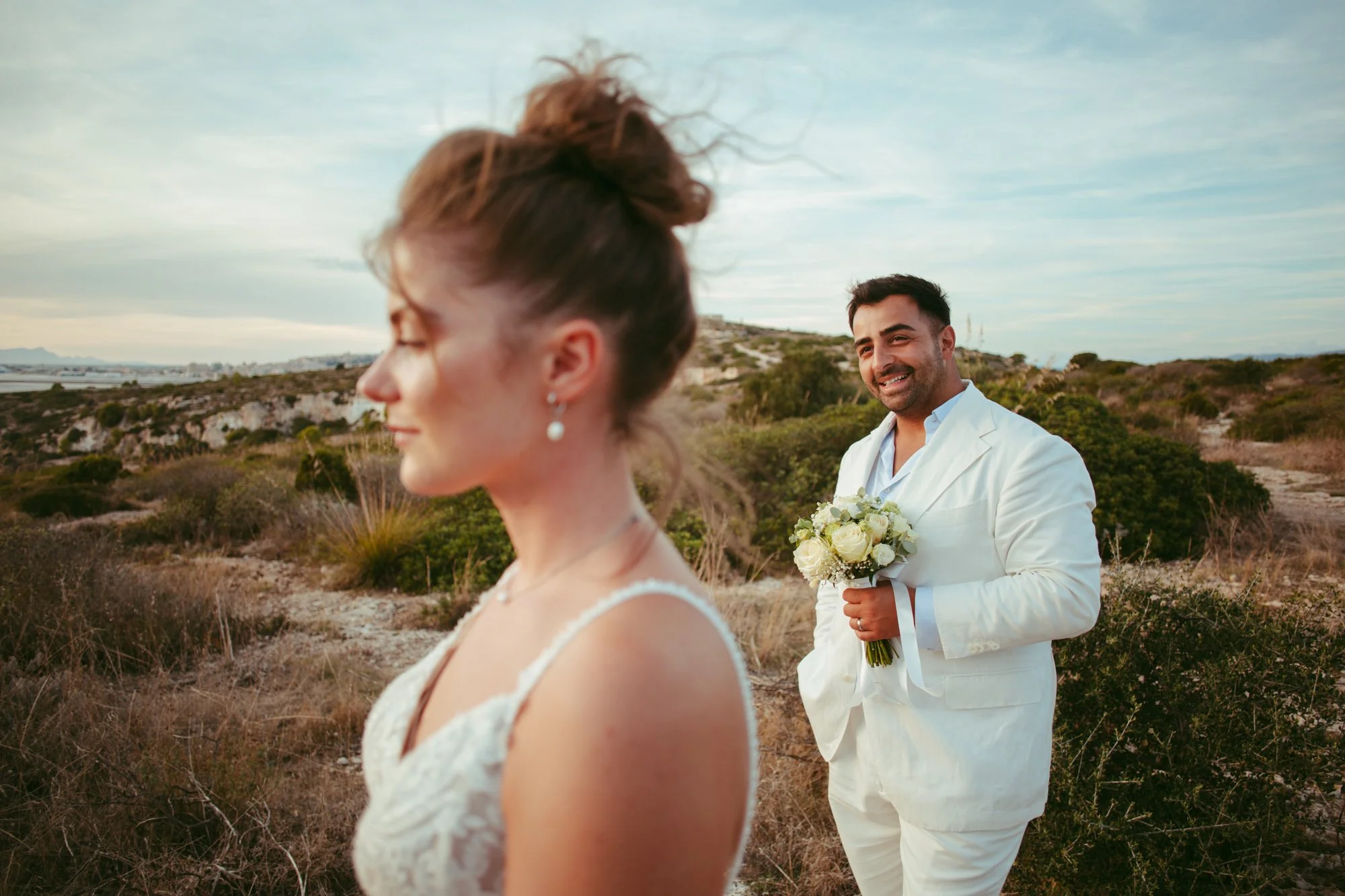 Primo piano dello sposo che guarda con dolcezza la fotocamera con il profilo sfocato della sposa in primo piano