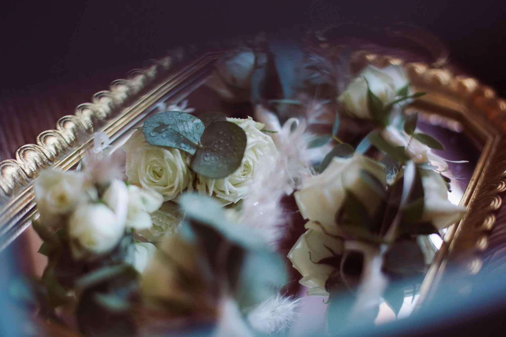 Bottoniere floreali con rose bianche ed eucalipto riflesse su uno specchio dorato, dettagli di lusso per lo sposo.