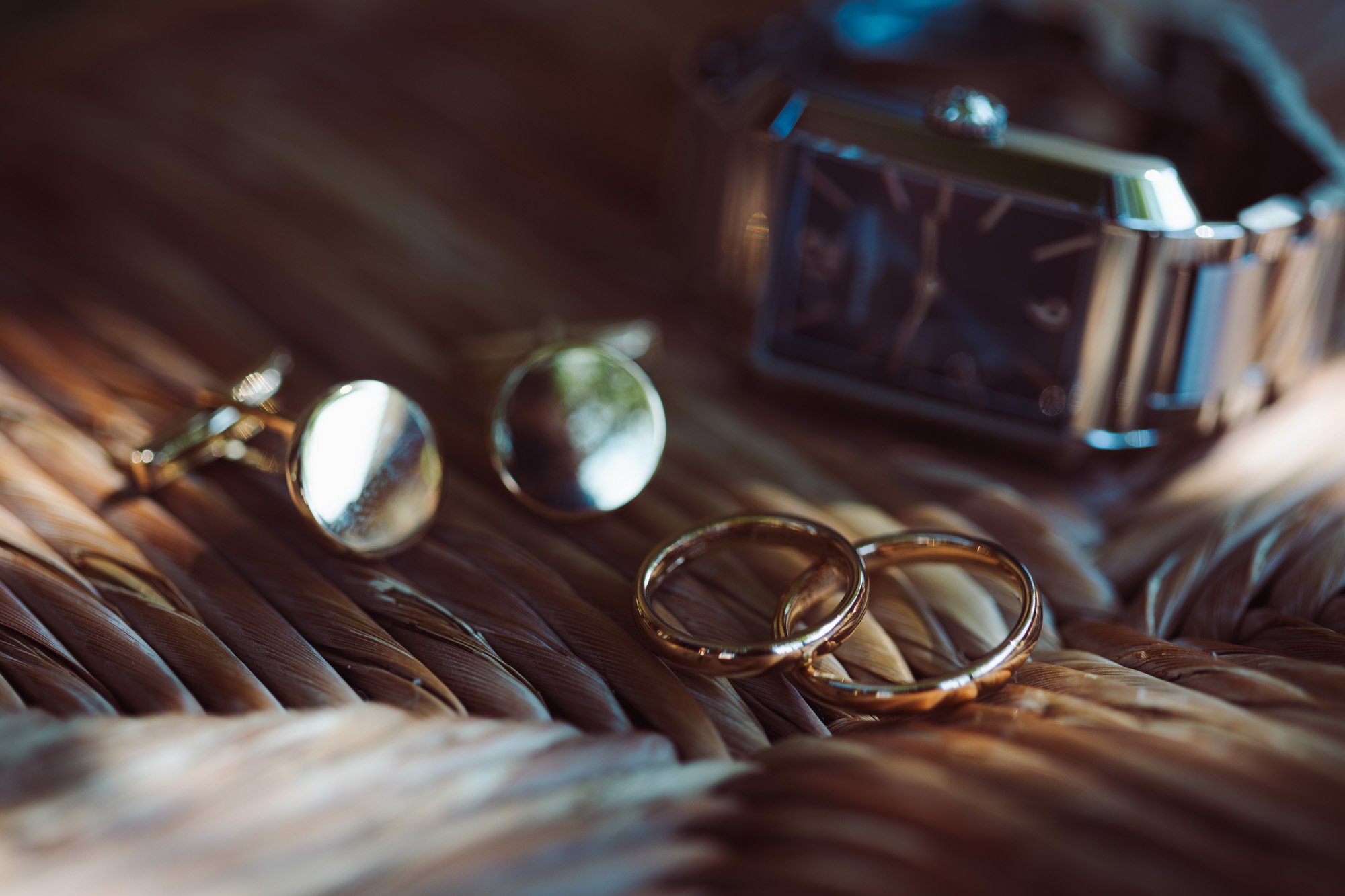 Dettagli di lusso dello sposo con fedi nuziali in oro, gemelli coordinati e orologio di design su fondo in vimini.