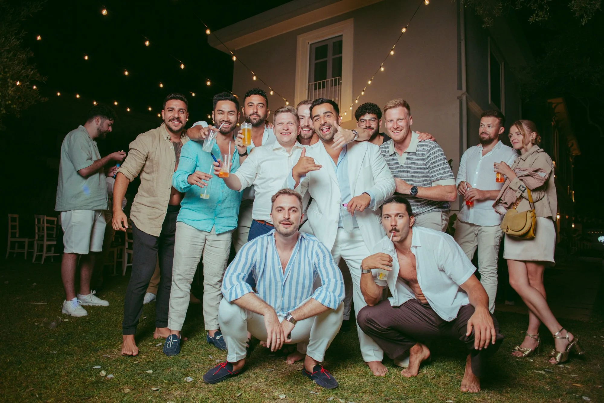 Lo sposo in abito bianco in posa divertente con il gruppo di amici con i drink in mano durante la festa serale