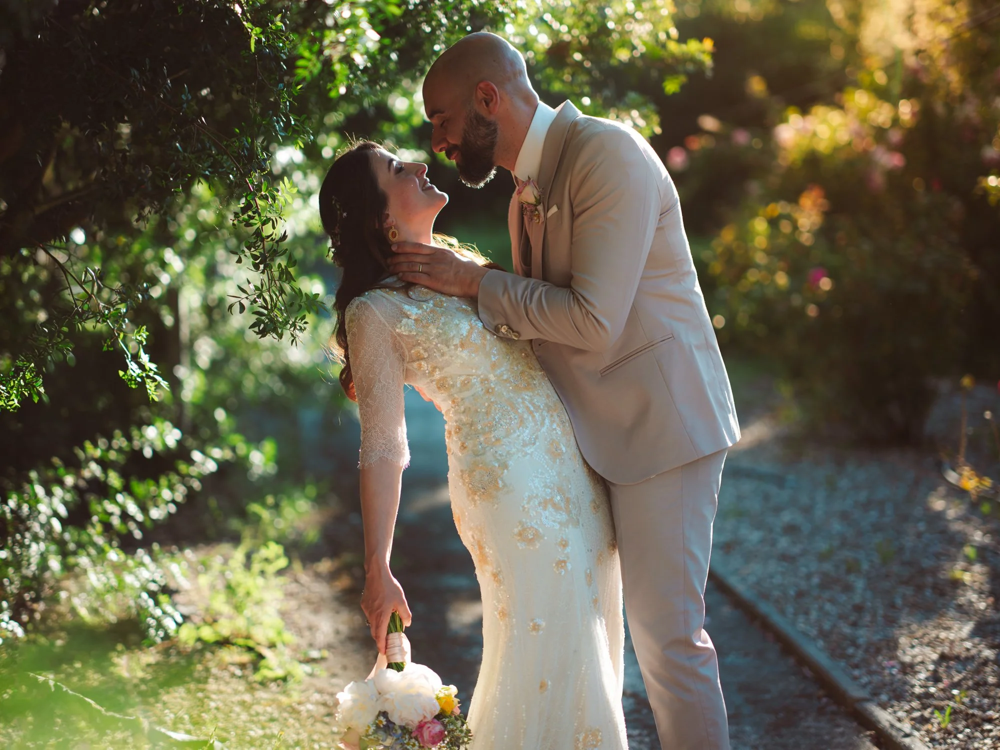 Sposo in abito beige che sostiene delicatamente la sposa in un elegante piegamento all'ombra degli alberi
