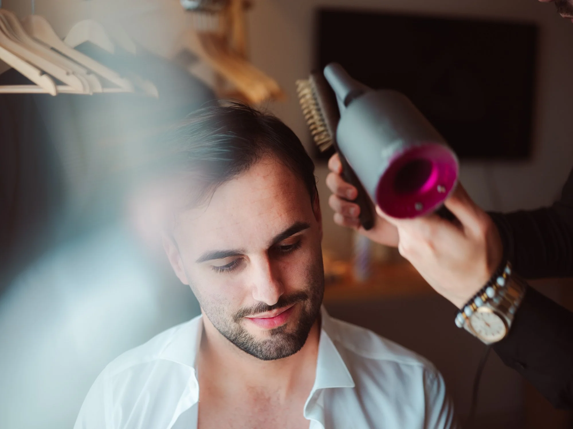 Primo piano dello sposo sorridente durante lo styling dei capelli con asciugacapelli prima della cerimonia