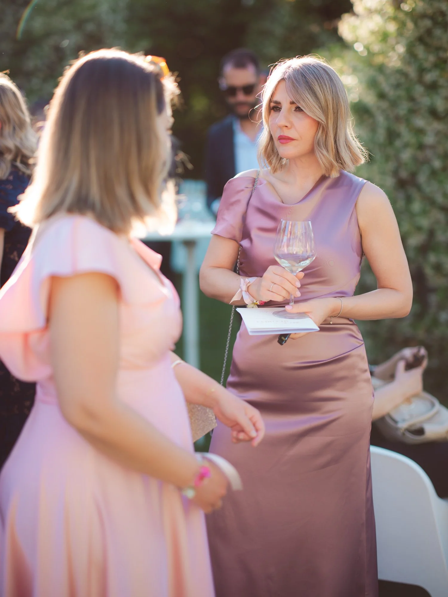 Invitata con taglio biondo e abito lucido rosa antico mentre conversa tenendo un calice di vino in mano