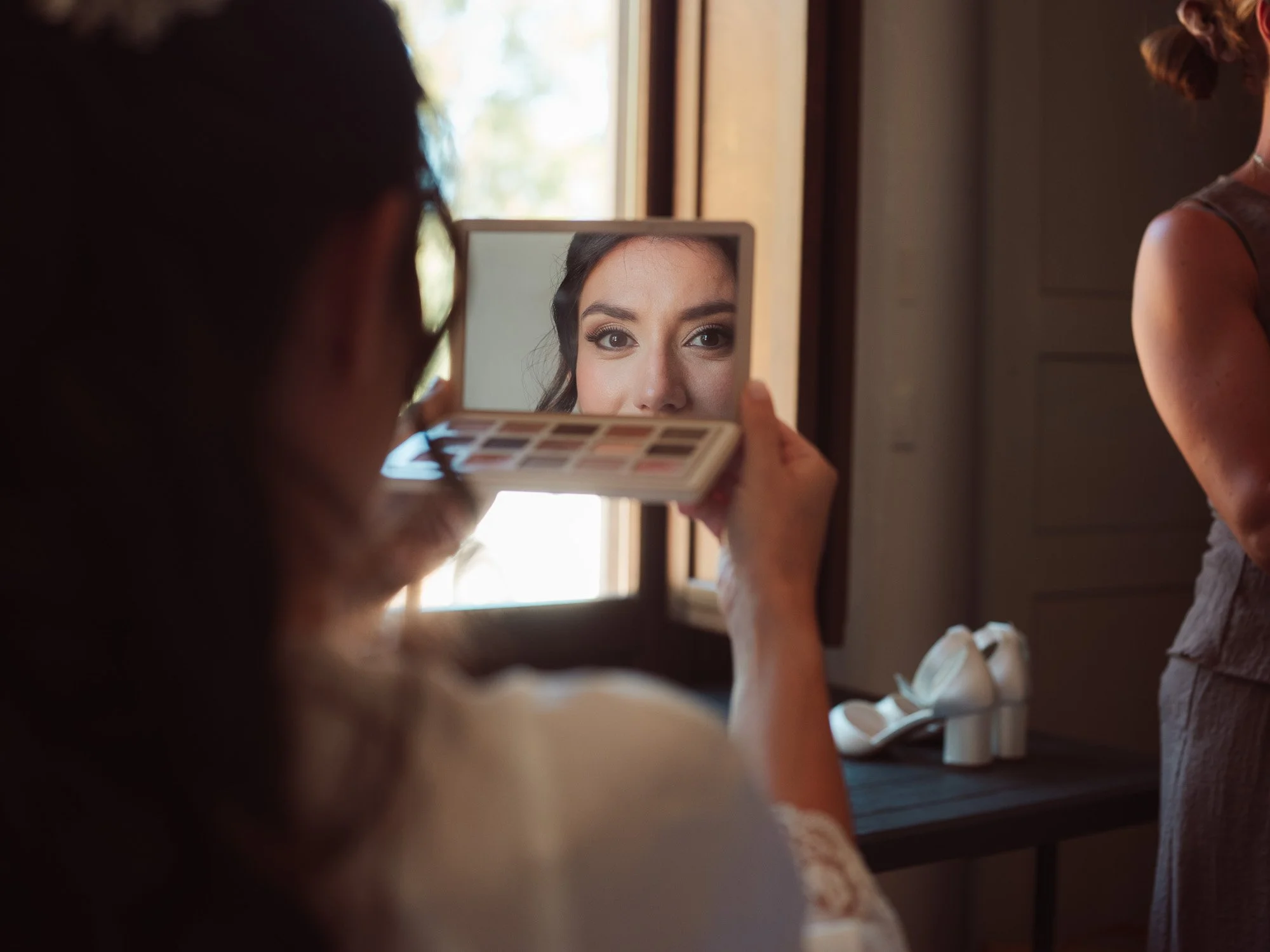 La sposa si specchia durante i preparativi nuziali in una stanza luminosa, con il trucco curato nei minimi dettagli.
