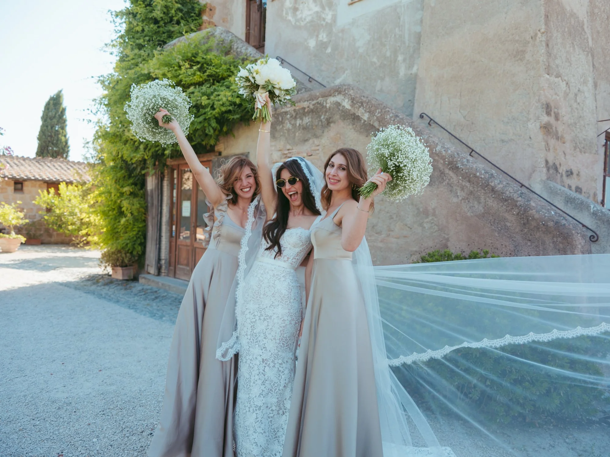 La sposa festeggia e sorride felice insieme alle sue due damigelle che sollevano i bouquet all'aperto