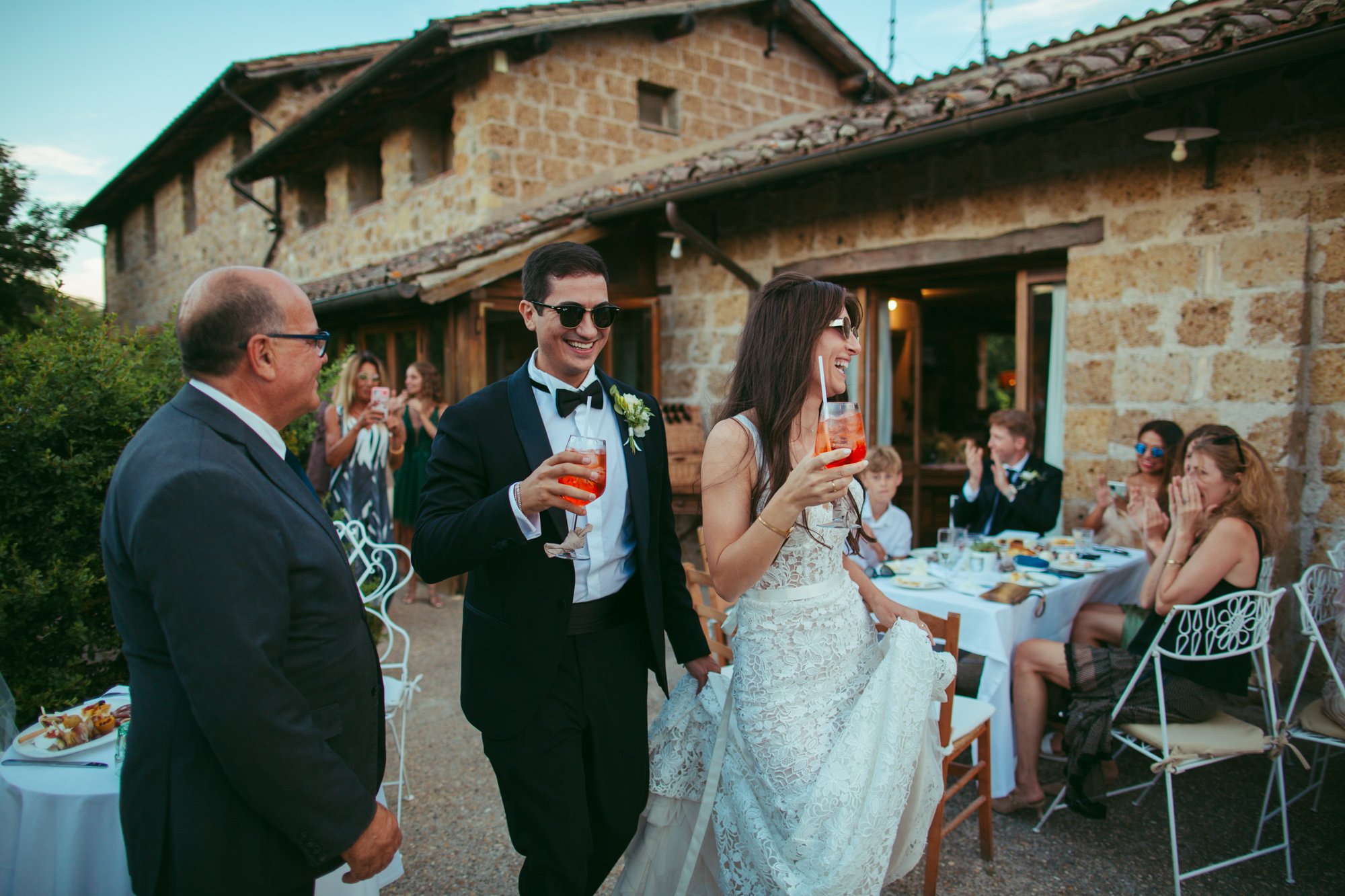 Gli sposi sorridenti con occhiali da sole brindano con uno spritz durante l'ingresso all'aperitivo all'aperto nel Borgo di Tragliata