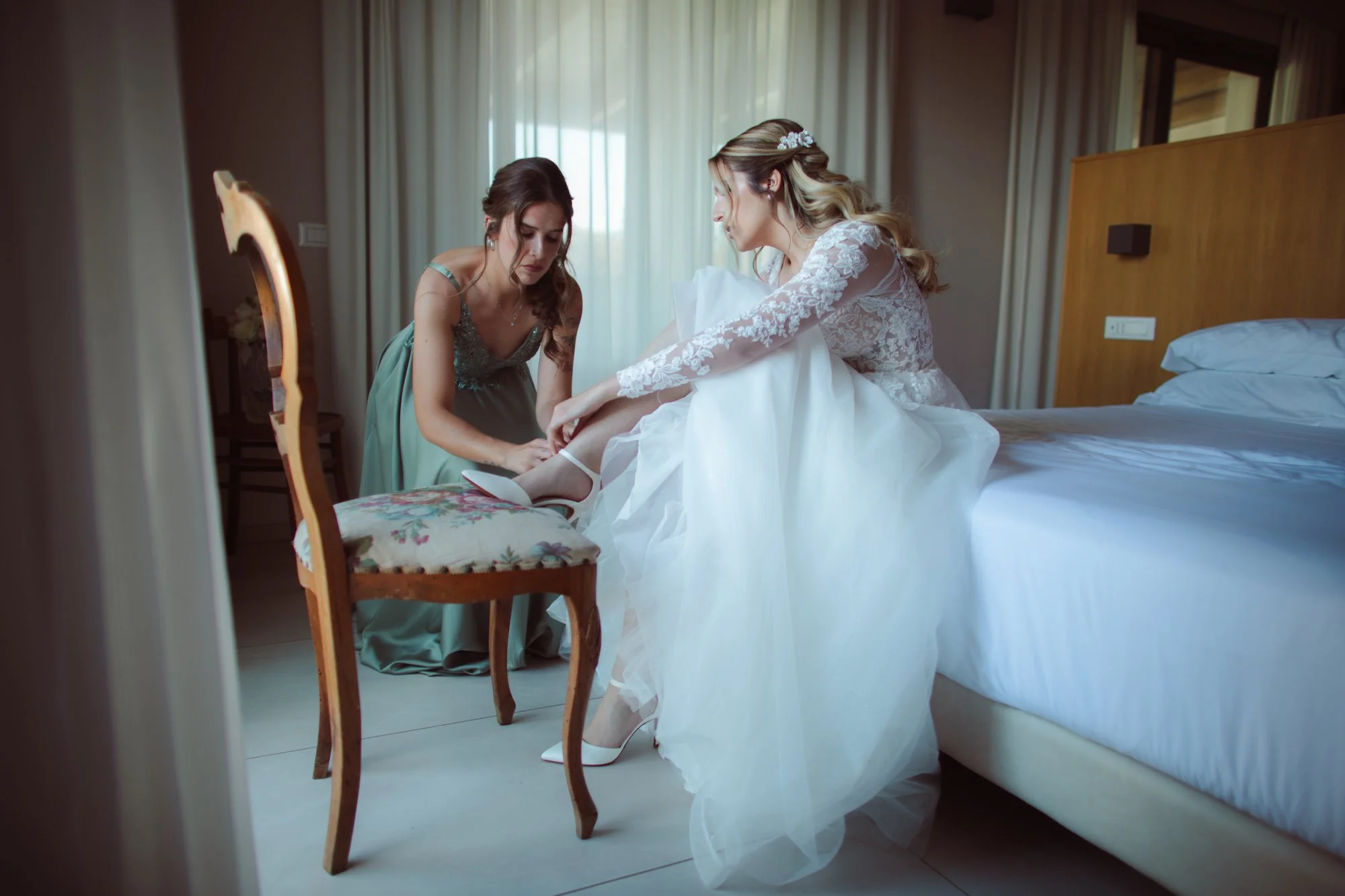 La sposa seduta sul letto in abito da sposa mentre un'amica in vestito verde la aiuta ad allacciare le scarpe