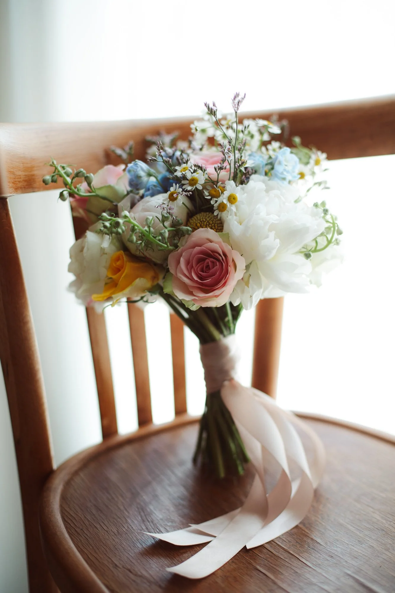 Bouquet da sposa con rose rosa e gialle, peonie bianche e margheritine appoggiato su una sedia in legno con nastro di seta
