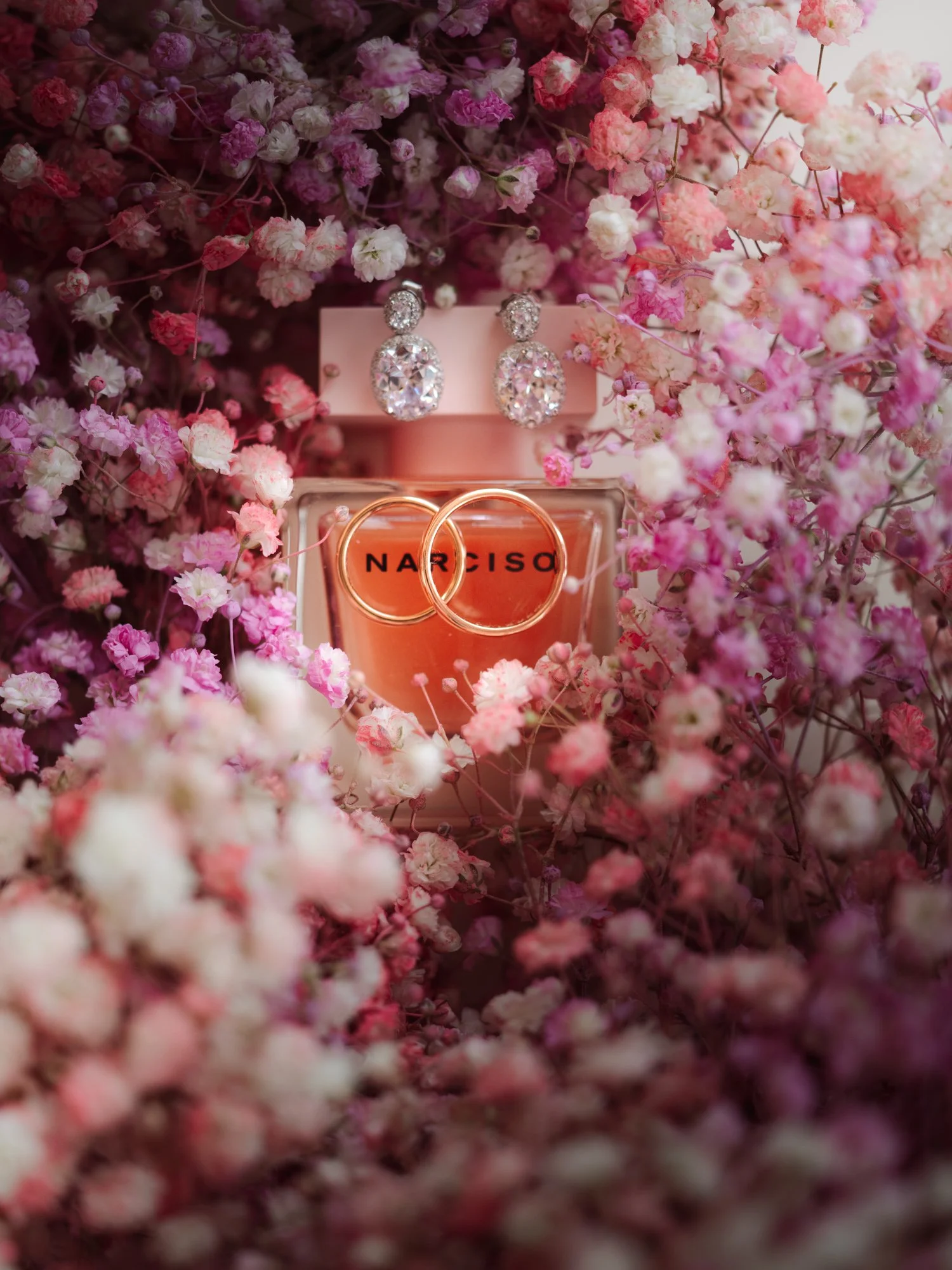 Orecchini di diamanti grandi e piccoli e fedi nuziali poggiati su una preziosa boccetta di profumo, circondati da fiori di vari colori rosa, bianco e viola.