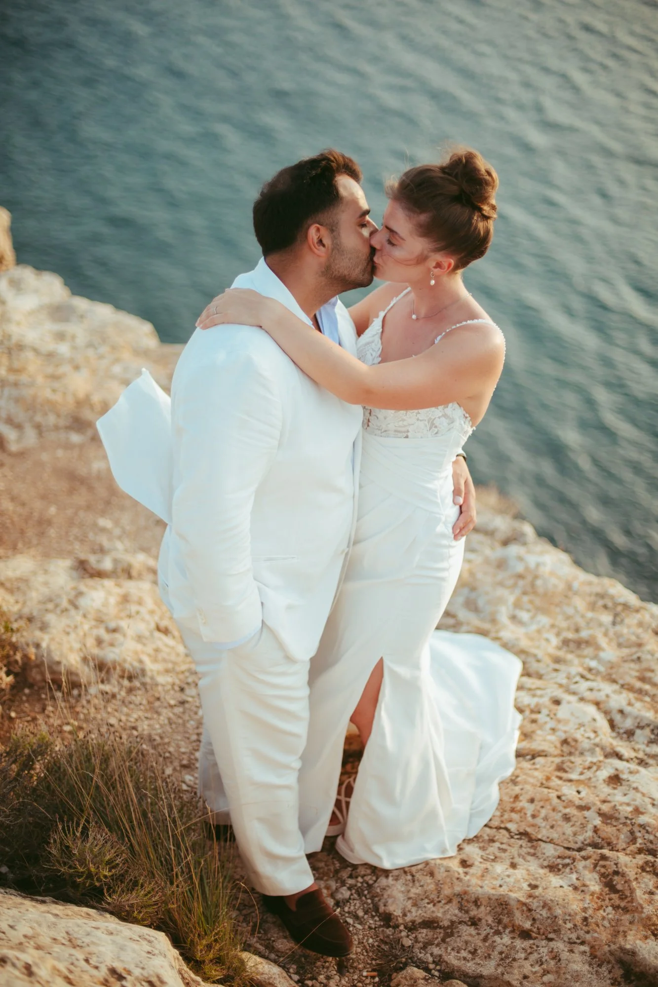 Gli sposi si scambiano un bacio appassionato sulla scogliera con il mare blu sullo sfondo