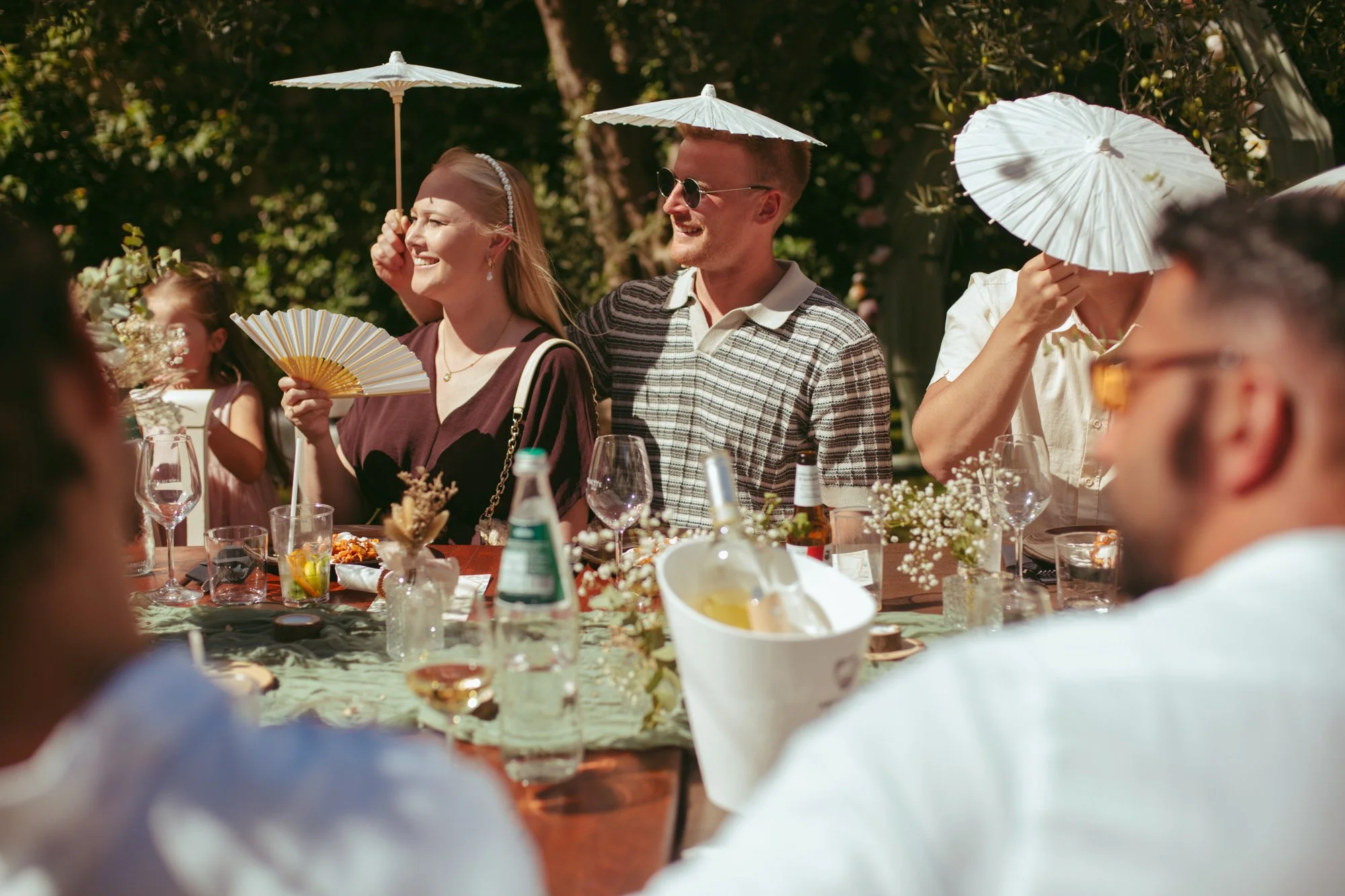 Invitati al tavolo che si riparano dal sole estivo utilizzando ombrellini bianchi in carta e ventagli