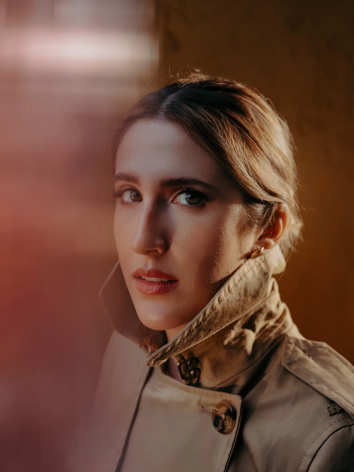 Primo piano editoriale di una donna in trench con luce calda di studio.