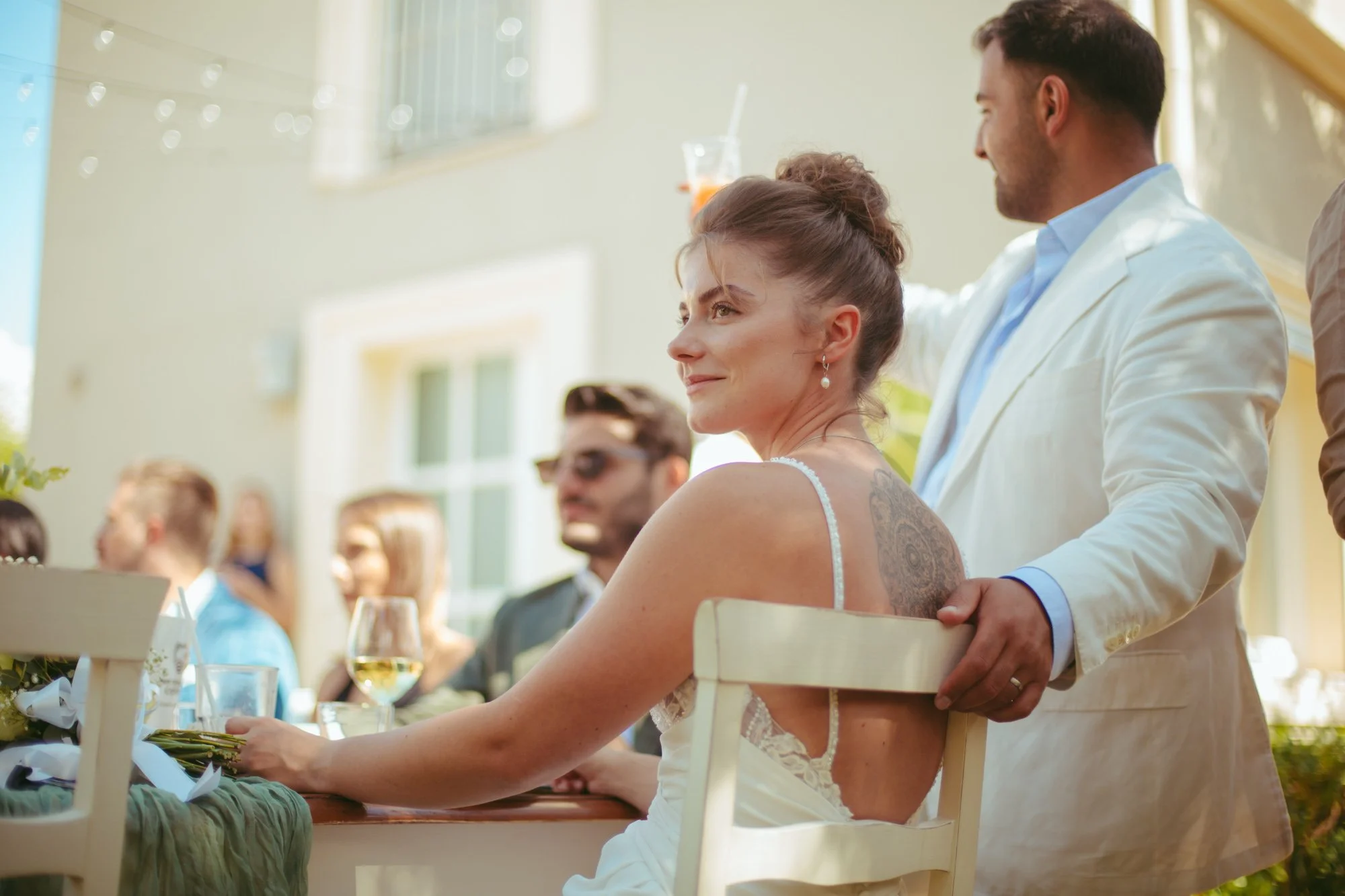 La sposa seduta al tavolo d'onore sorride mentre lo sposo in abito bianco le appoggia dolcemente la mano sulla sedia durante il pranzo di nozze