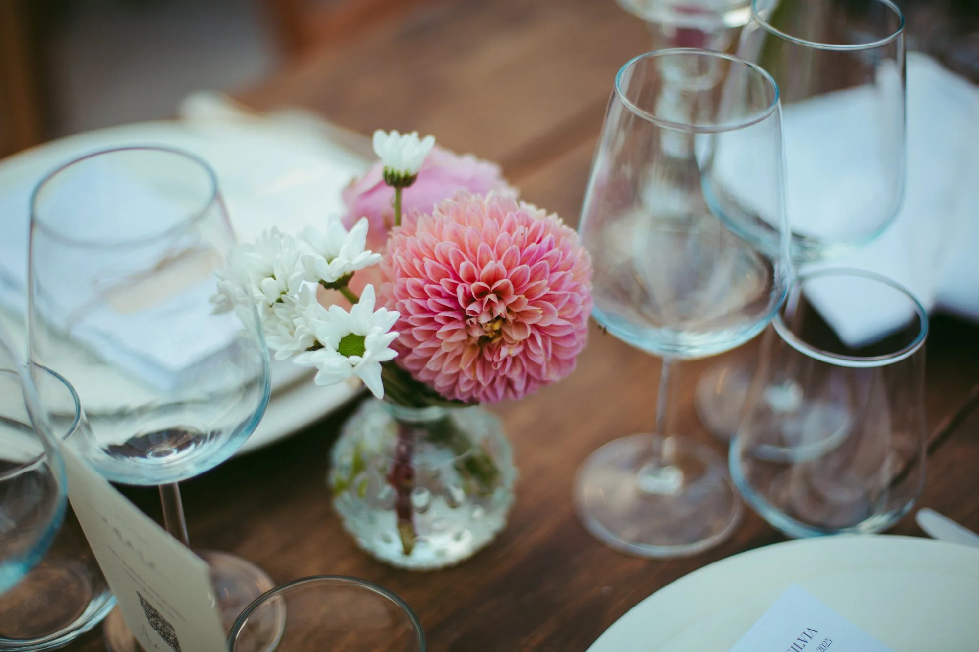 Dettaglio ravvicinato della mise en place con vaso in vetro, dalie rosa e margherite per il ricevimento a Tragliata