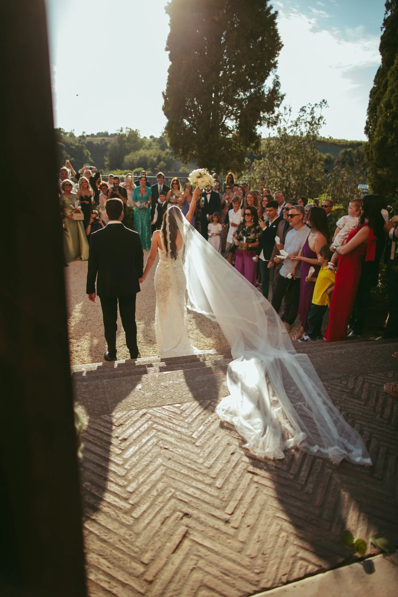 La sposa solleva il bouquet in segno di gioia mentre scende la scalinata mano nella mano con lo sposo verso la folla di invitati