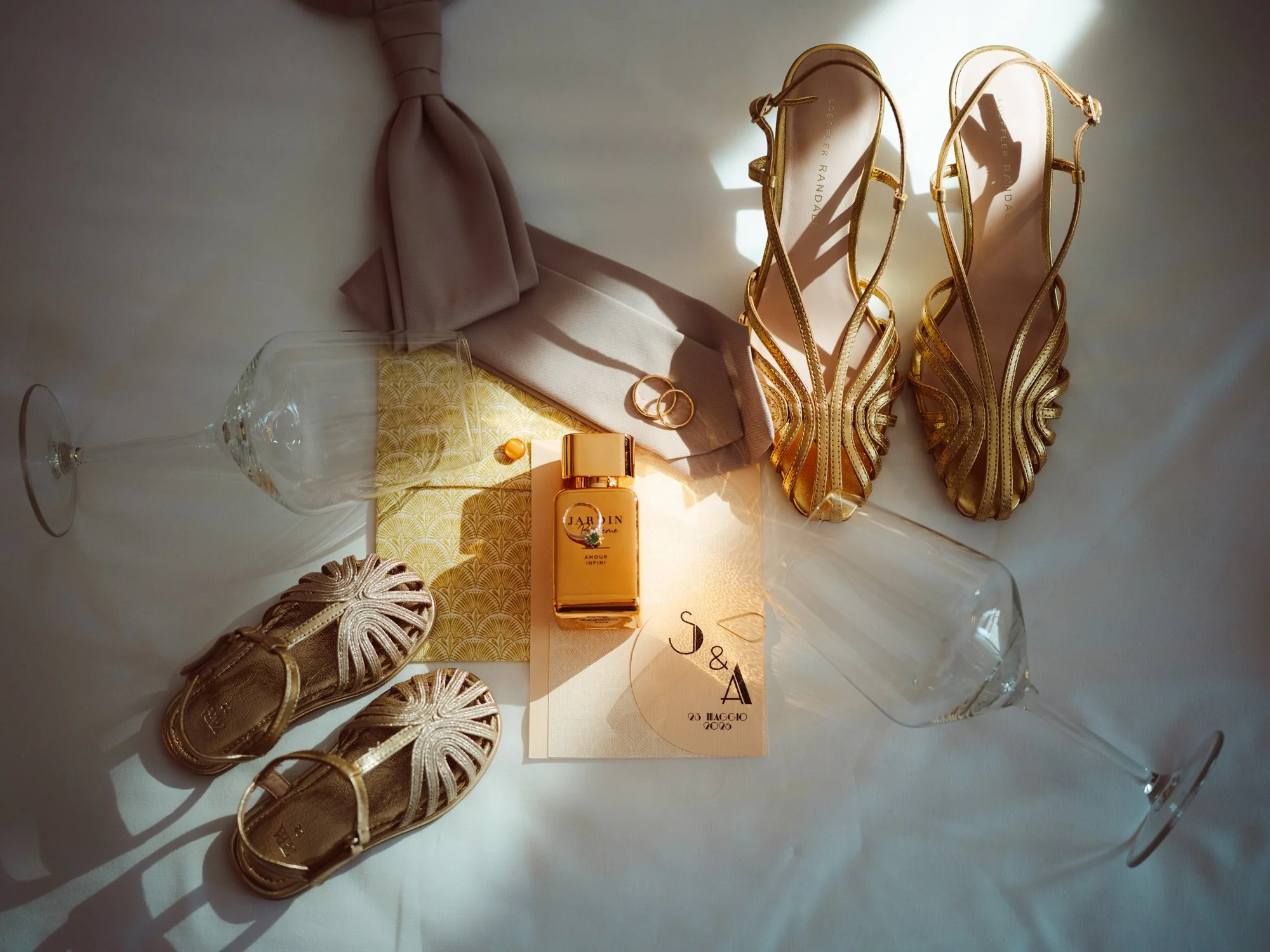 Composizione flat lay di accessori matrimoniali con scarpe dorate da donna, sandaletti da bimba, cravatta, flacone di profumo e fedi su partecipazioni