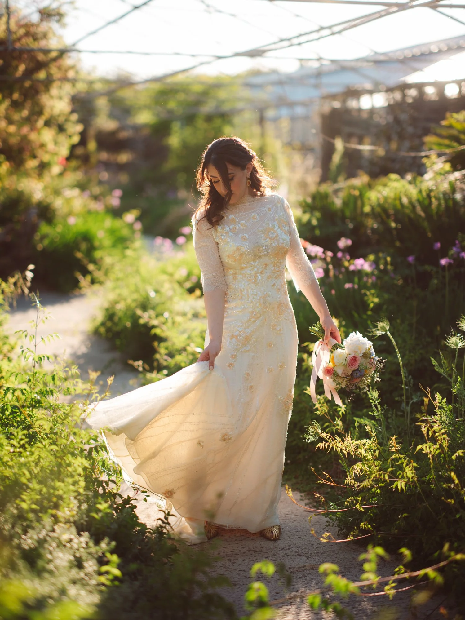 Sposa a figura intera mentre solleva delicatamente il lembo dell'abito con ricami dorati tenendo il bouquet nell'altra mano