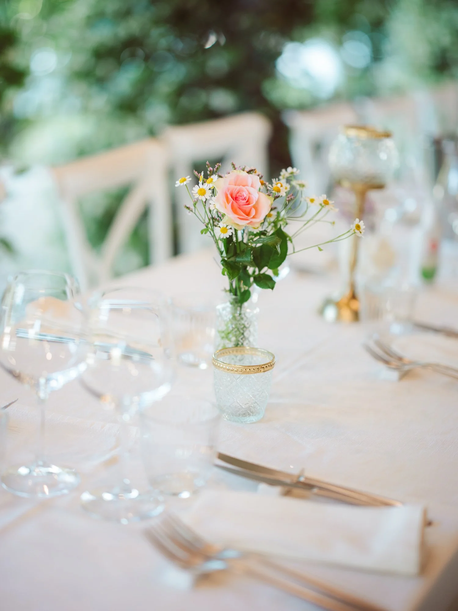 Mise en place elegante su tovaglia bianca con bicchieri di cristallo e centrotavola con rosa rosa e margherite