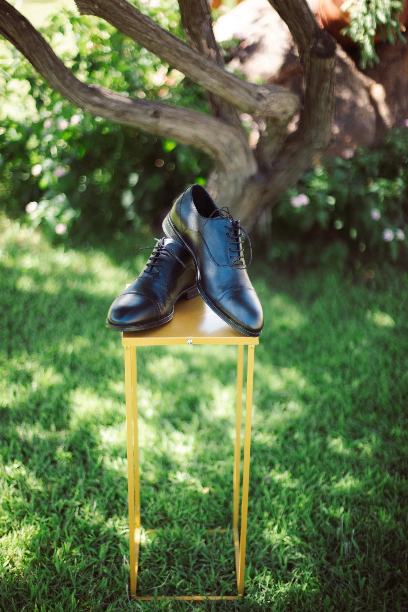 Scarpe classiche stringate in pelle nera da sposo posizionate su un elegante supporto dorato all'aperto.
