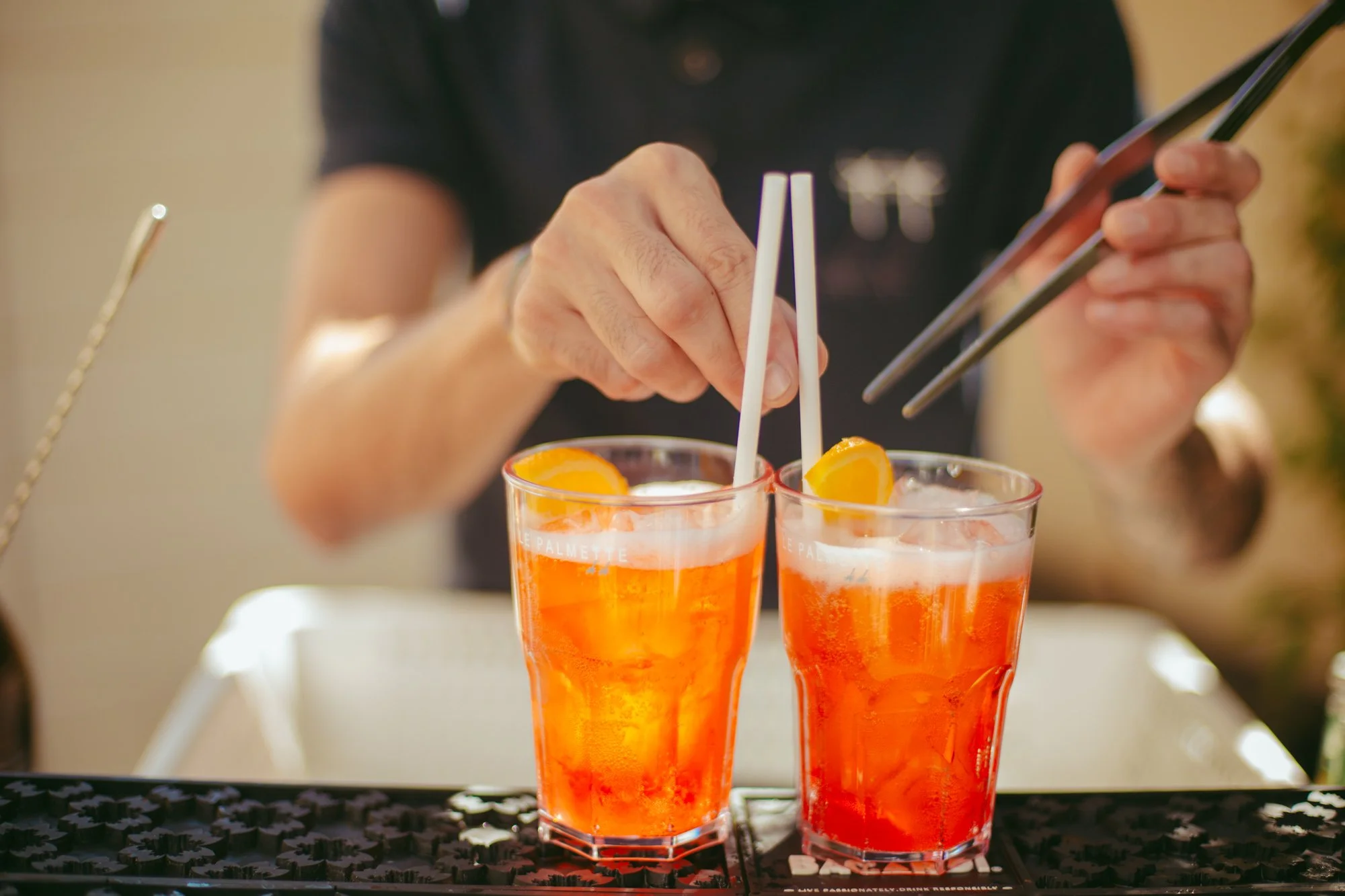Primo piano delle mani del bartender che guarnisce due bicchieri di Aperol Spritz con cannucce e fettine d'arancia
