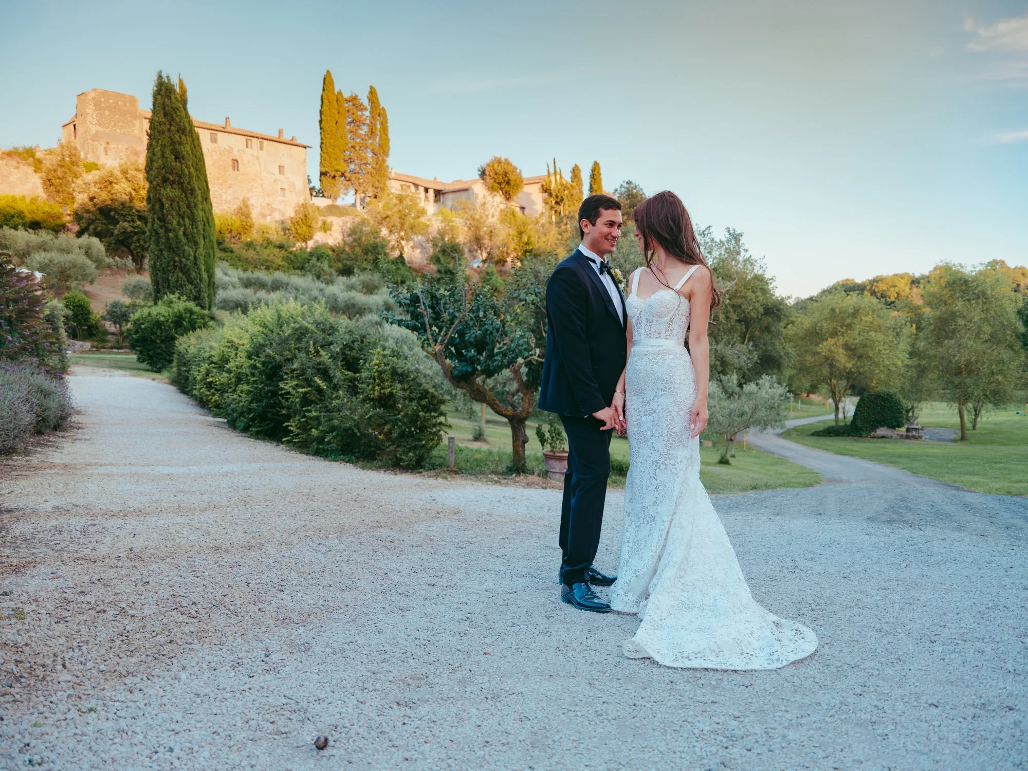 Ritratto a figura intera degli sposi su un viale in ghiaia con la sposa nel suo abito in pizzo e uno storico edificio del Borgo di Tragliata sullo sfondo