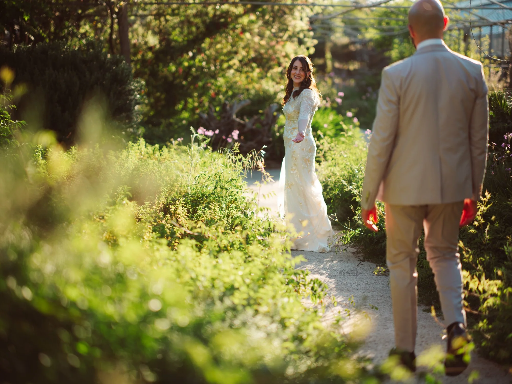 Sposa sorridente in abito con pizzo e paillettes dorate si volta e tende la mano verso lo sposo su un viale alberato