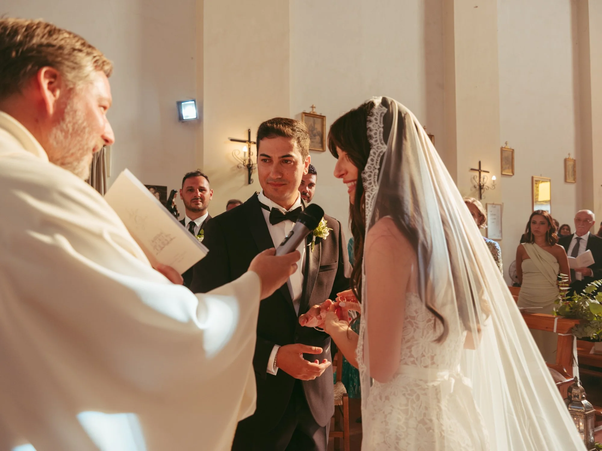 La sposa infila la fede nuziale allo sposo con il sacerdote che sostiene il microfono durante la cerimonia