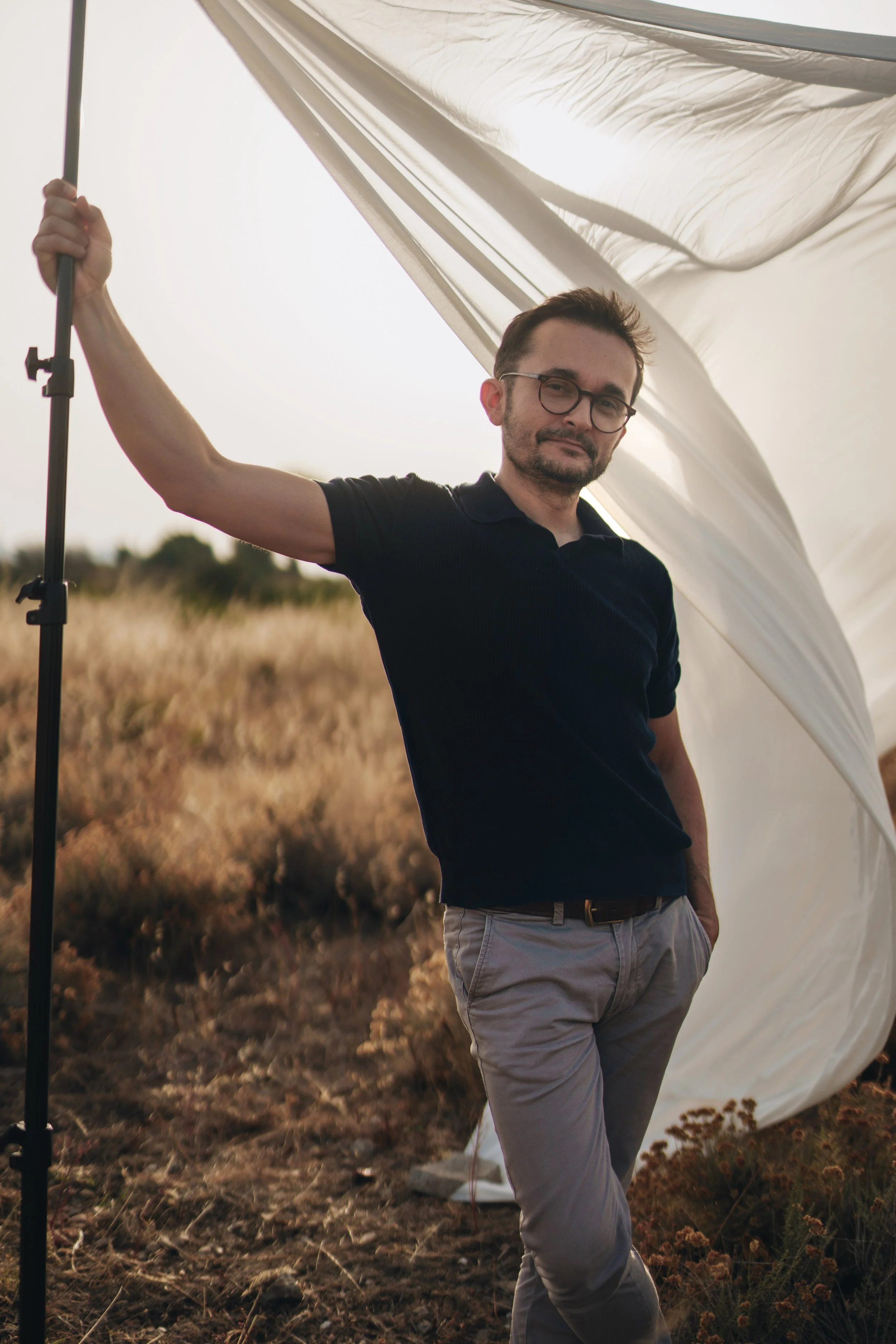 Uomo con occhiali, in piedi in un campo, si regge a una leggera struttura a cui è appeso un morbido e leggero telo bianco mosso dal vento. È vestito con una polo in maglia blu scuro e pantaloni beige.