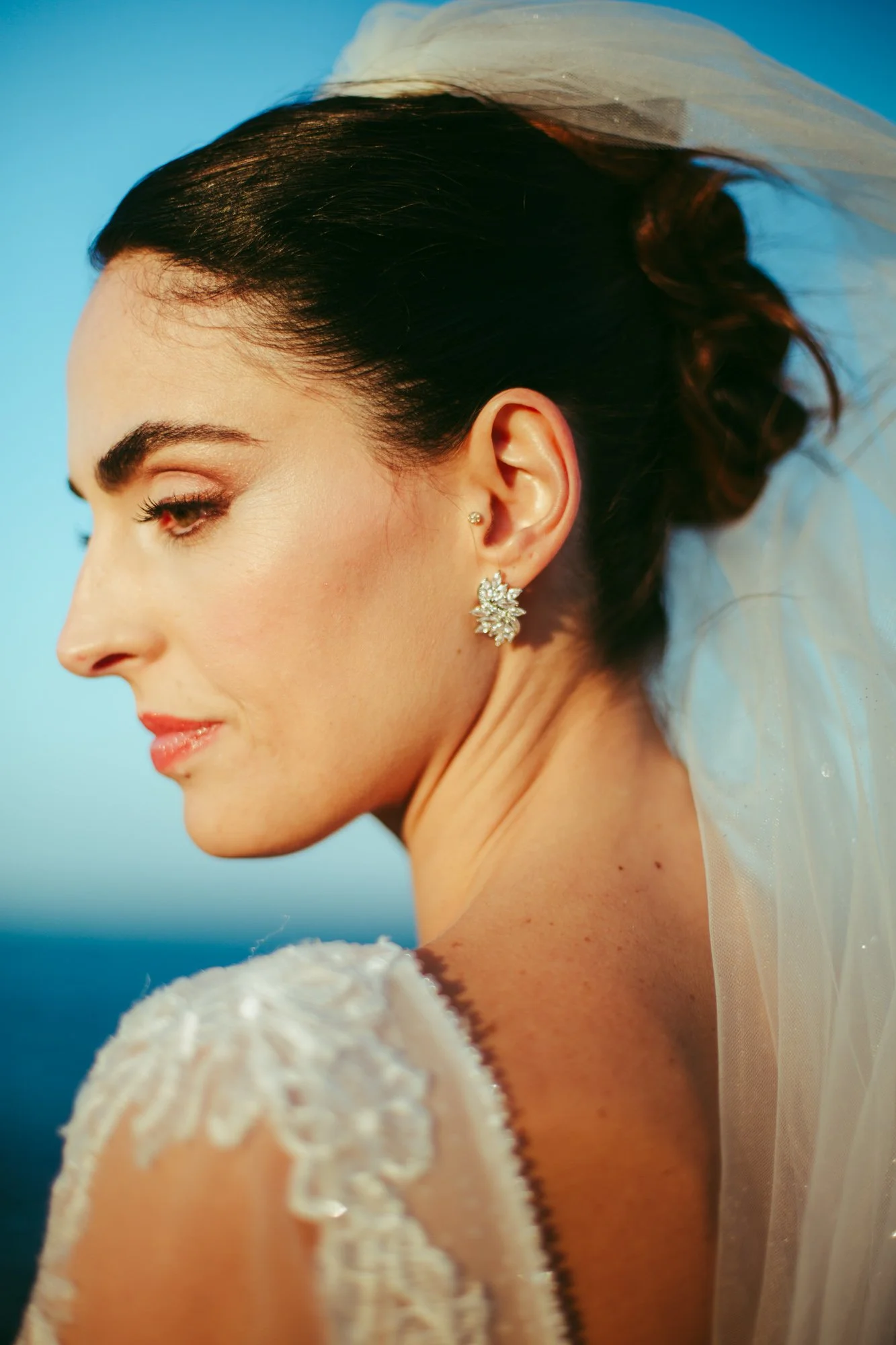 Primo piano di una sposa con orecchini gioiello, capelli scuri acconciati, abito bianco ricamato, con uno sfondo blu. La sua espressione è serena.