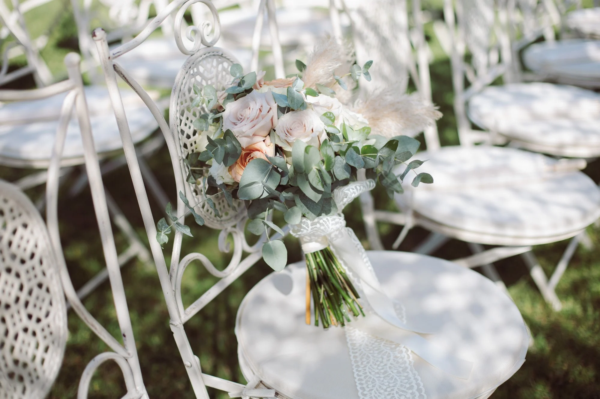 Elegante bouquet da sposa in toni pastello poggiato su una sedia in ferro battuto bianco tra i giochi di luce del giardino.