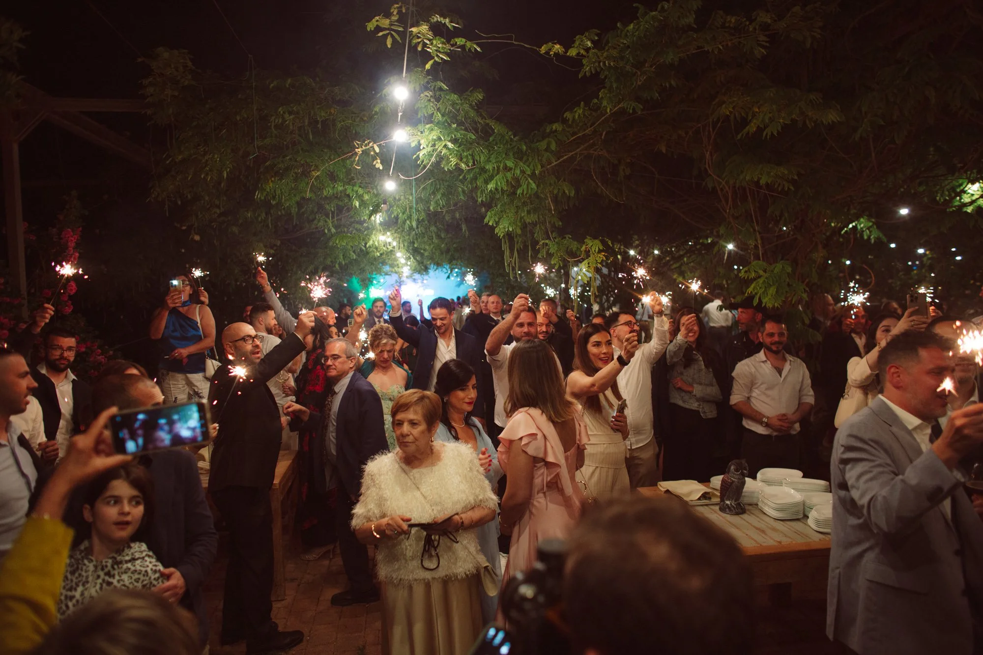Panoramica notturna del giardino con tutti gli invitati che alzano le stelline luminose accese sotto le catene di luci
