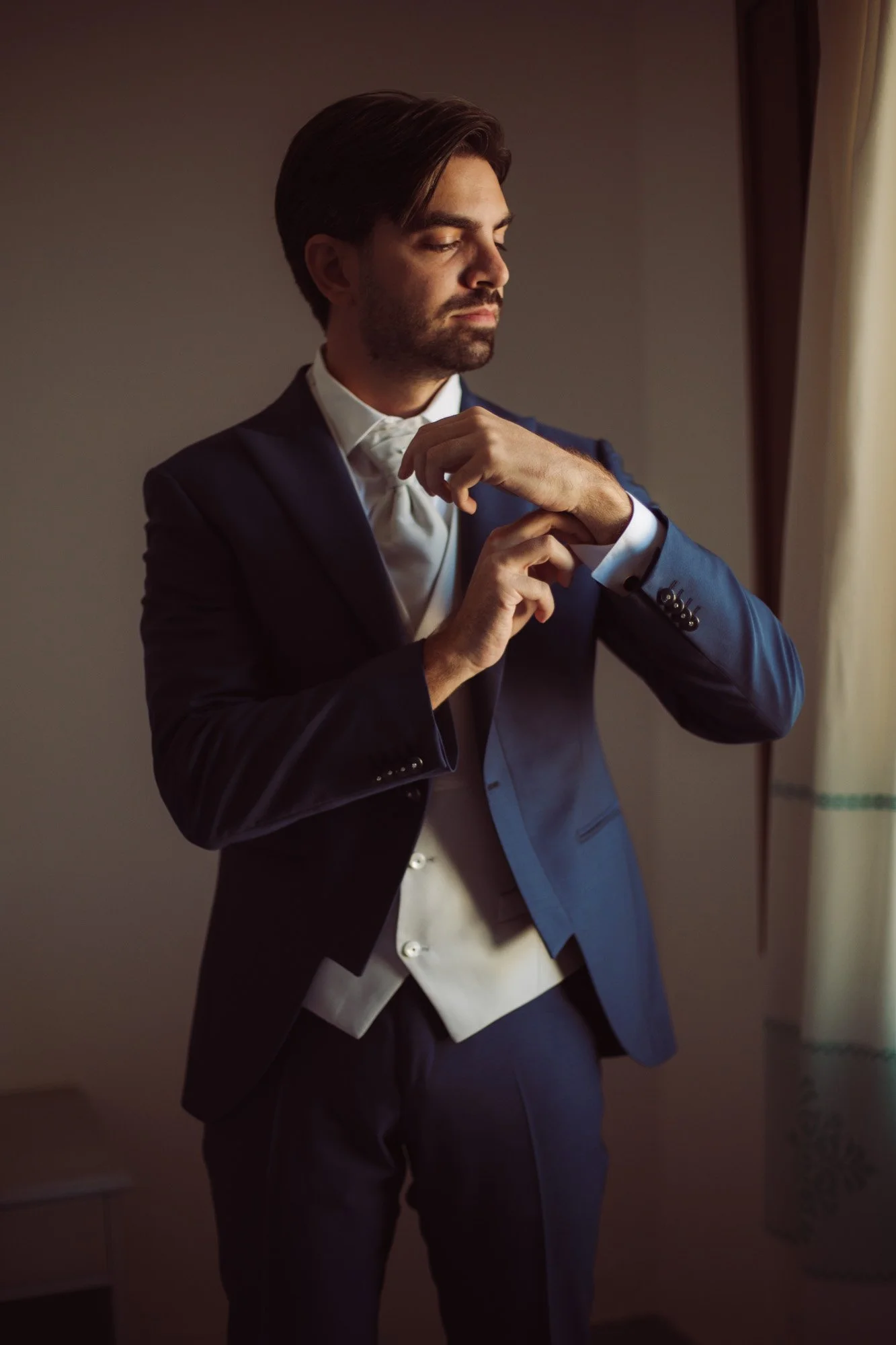 Lo sposo in abito blu e gilet chiaro si sistema i polsini della camicia alla luce della finestra prima della cerimonia.