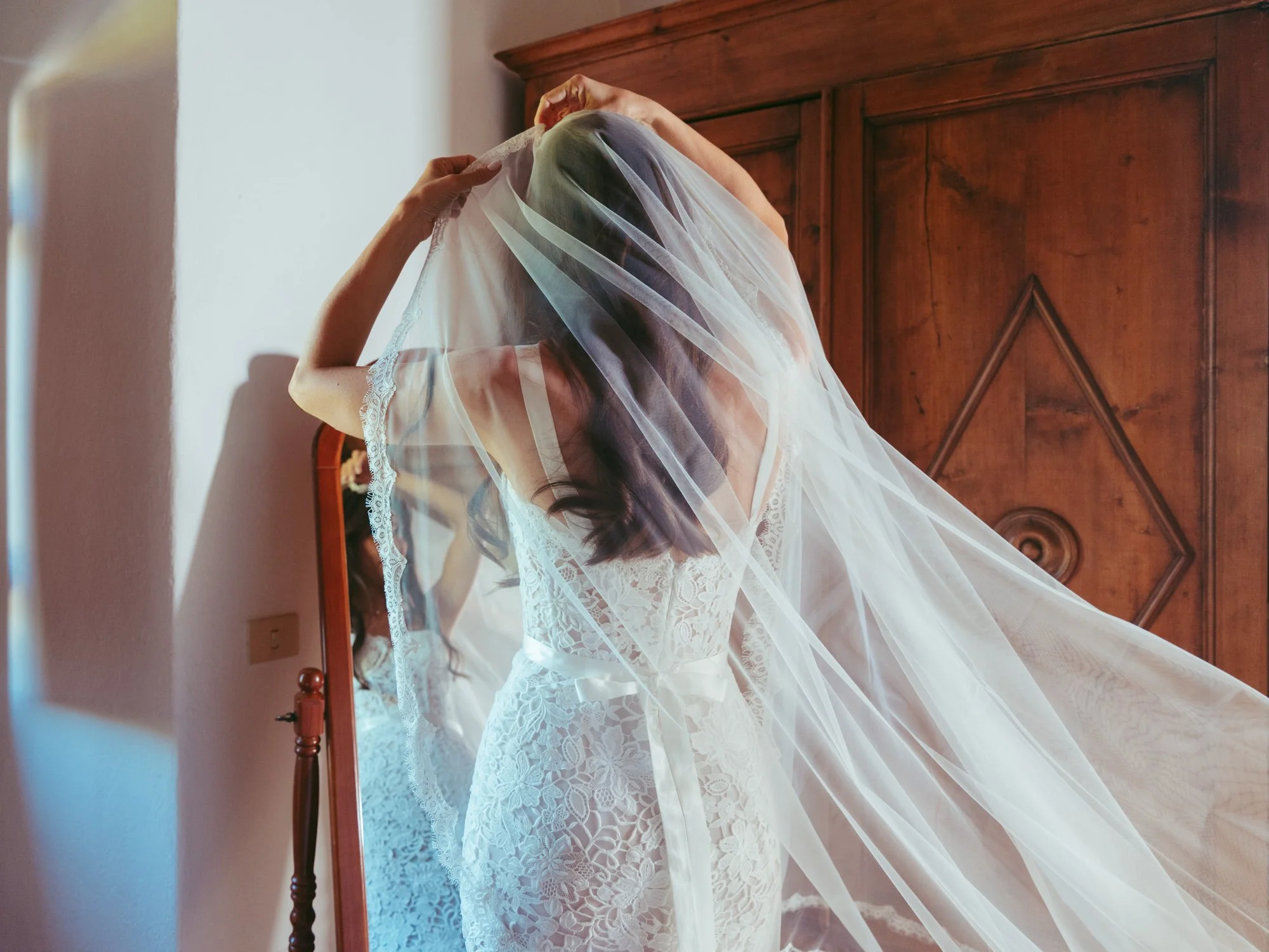 Sposa di spalle davanti allo specchio mentre indossa il lungo velo bordato in pizzo