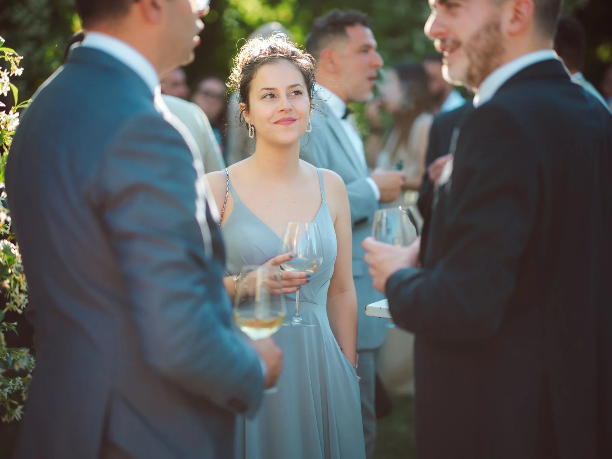 Invitata sorridente in abito azzurro spalline sottili mentre parla con altri ospiti tenendo un calice di vino in mano durante l'aperitivo