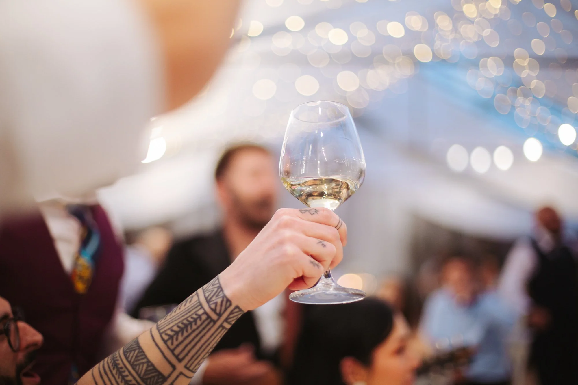 Primo piano di una mano con tatuaggi geometrici che solleva un calice di vino bianco durante il ricevimento