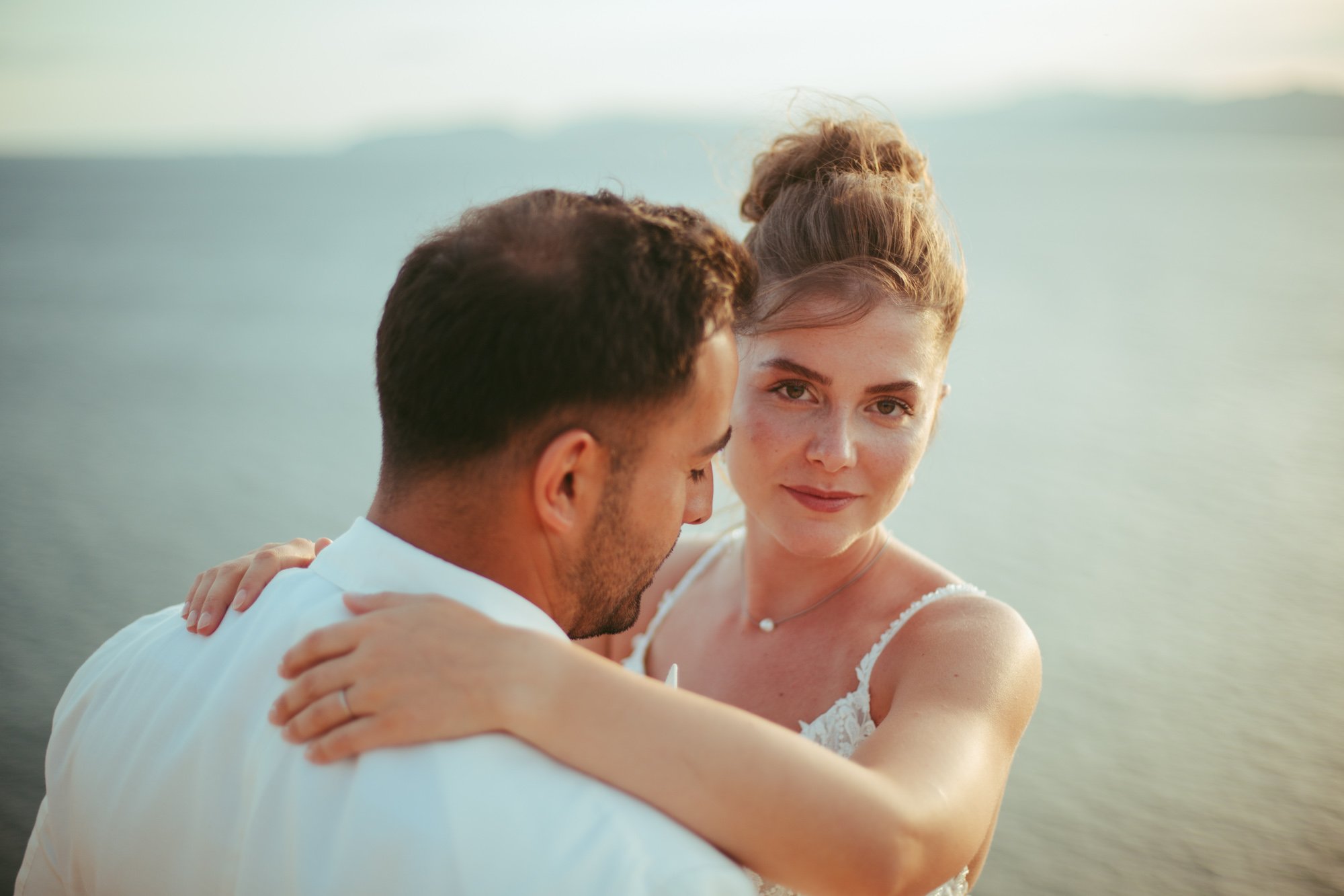 La sposa guarda la fotocamera mentre abbraccia lo sposo da dietro sulla costa al tramonto