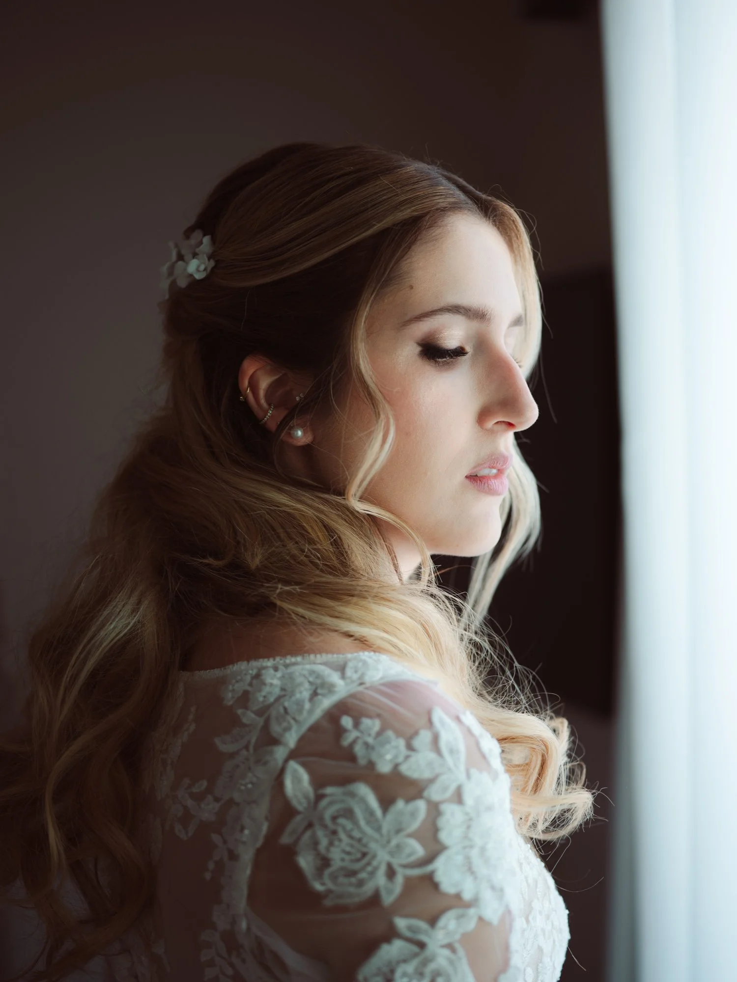 Primo piano di profilo della sposa con acconciatura semiraccolta, fermaglio floreale e abito in pizzo illuminata dalla luce della finestra